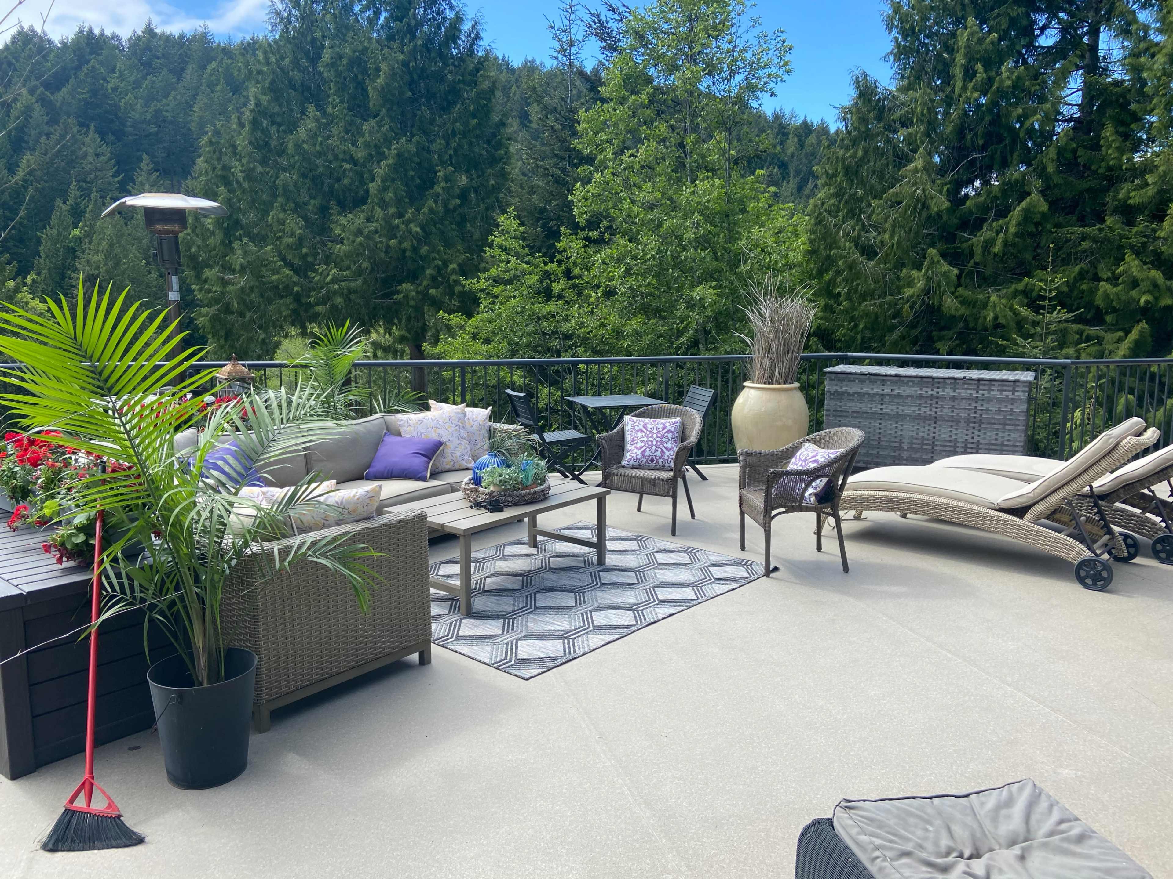 A patio area featuring a seating arrangement with a table, a chaise lounge, planters, and a view of trees in the background.