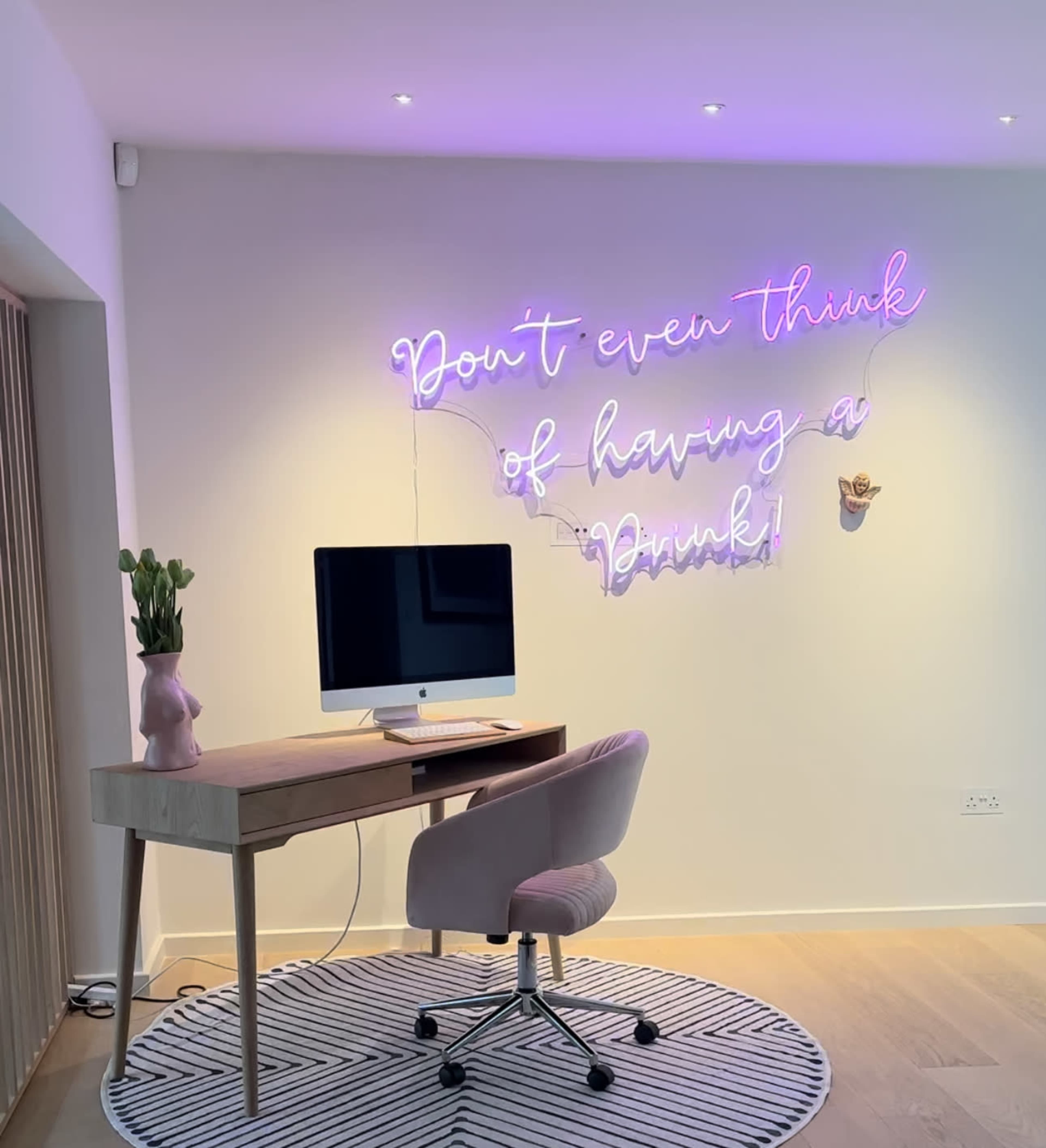 A modern workspace features a wooden desk with an iMac, a pink chair, and a neon sign on the wall that reads, "Don't even think of having a drink!"