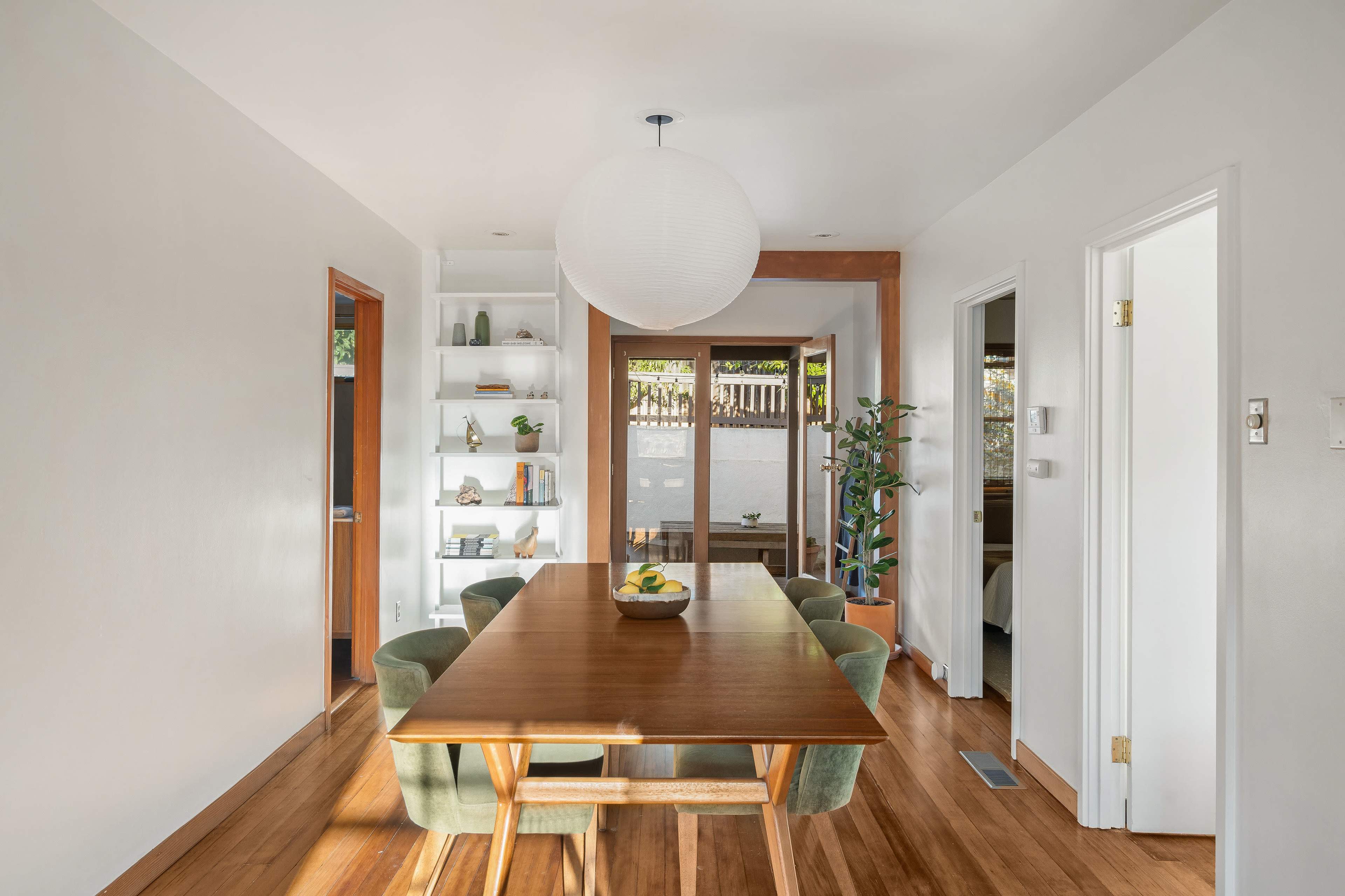 A dining area features a wooden table surrounded by green chairs, with a large pendant light overhead and access to a patio through sliding glass doors.