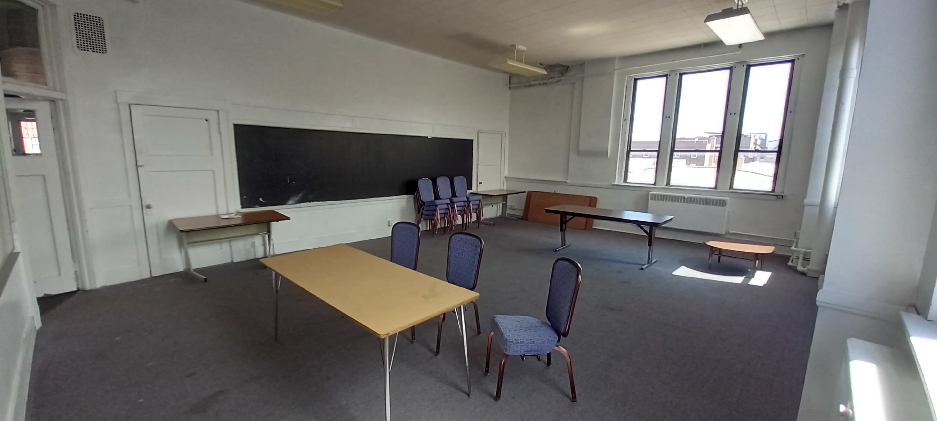 An empty classroom features a blackboard, several chairs stacked against the wall, and a few tables arranged with ample open space.