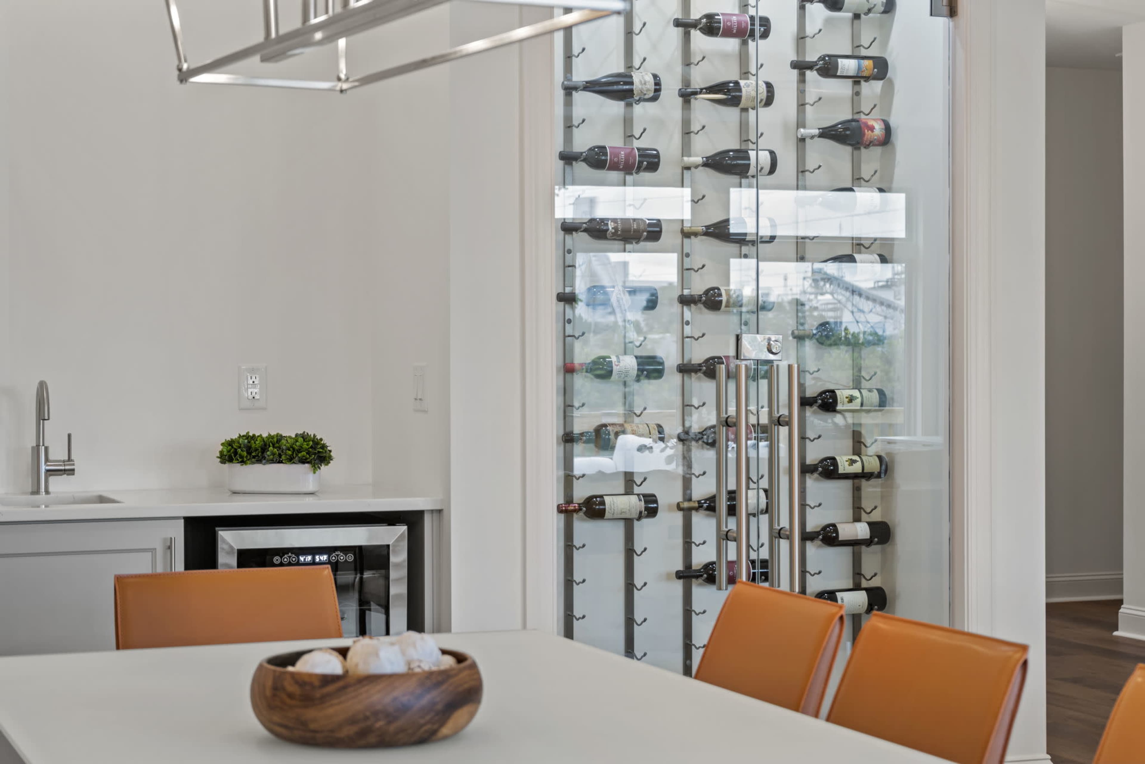 A modern kitchen features a wine rack displaying several bottles along a glass wall next to a dining table with brown leather chairs and a wooden bowl.