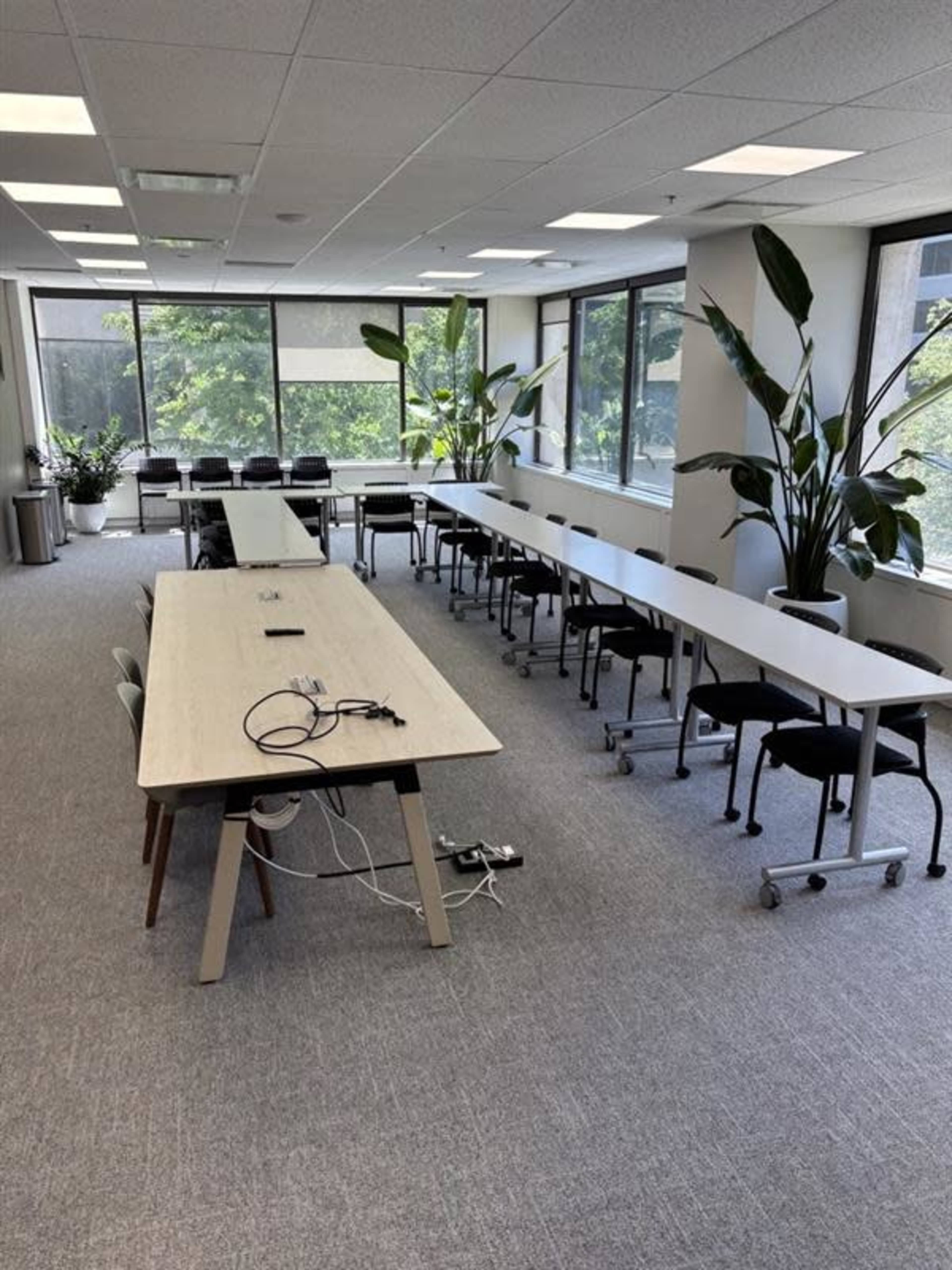 The image shows a modern conference room with two long tables arranged in a U-shape, surrounded by chairs and large windows allowing natural light to enter.