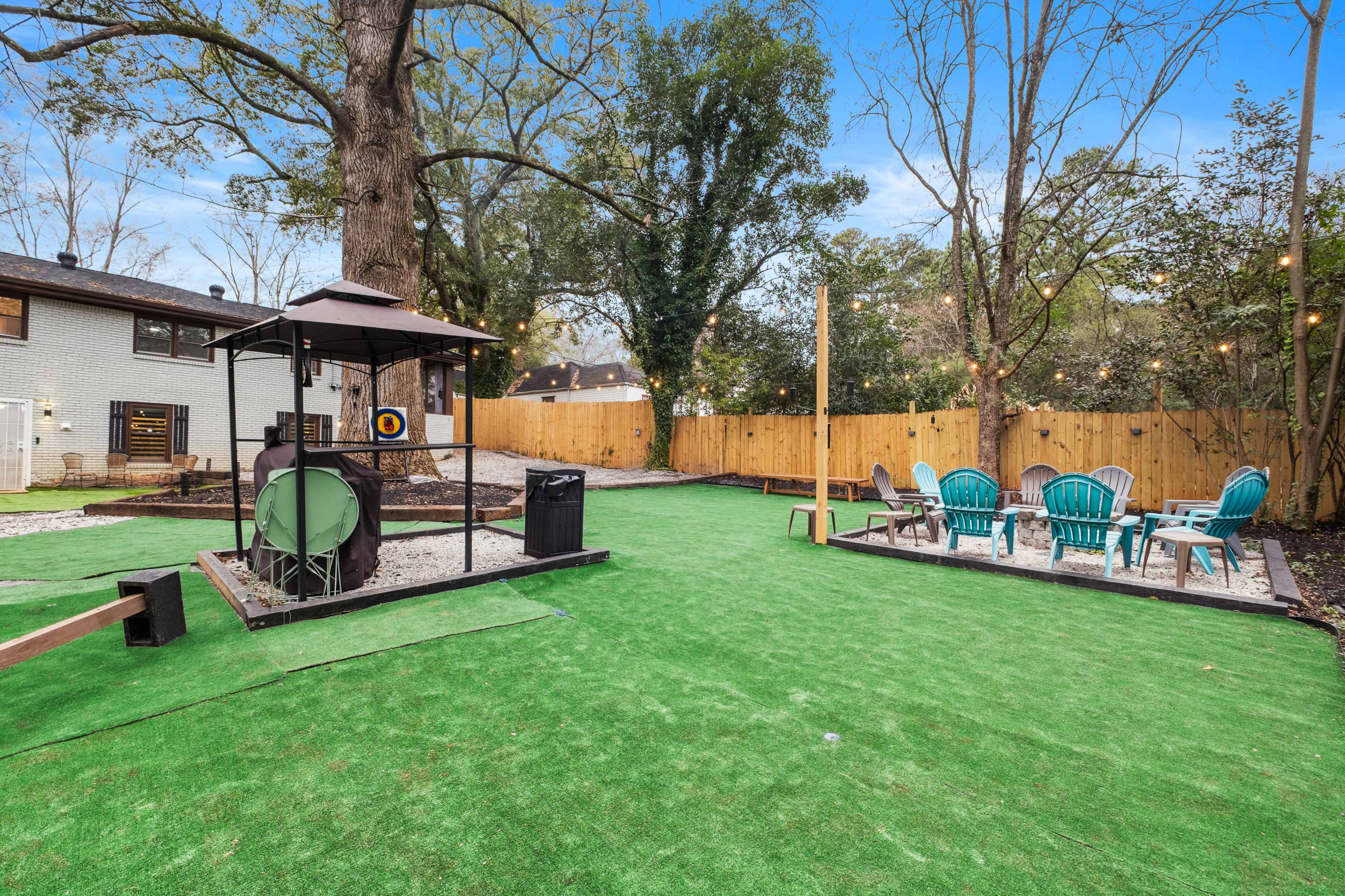 The image shows a backyard with artificial turf, a gazebo with a target, a trash can, and a dining area featuring a table and blue chairs.
