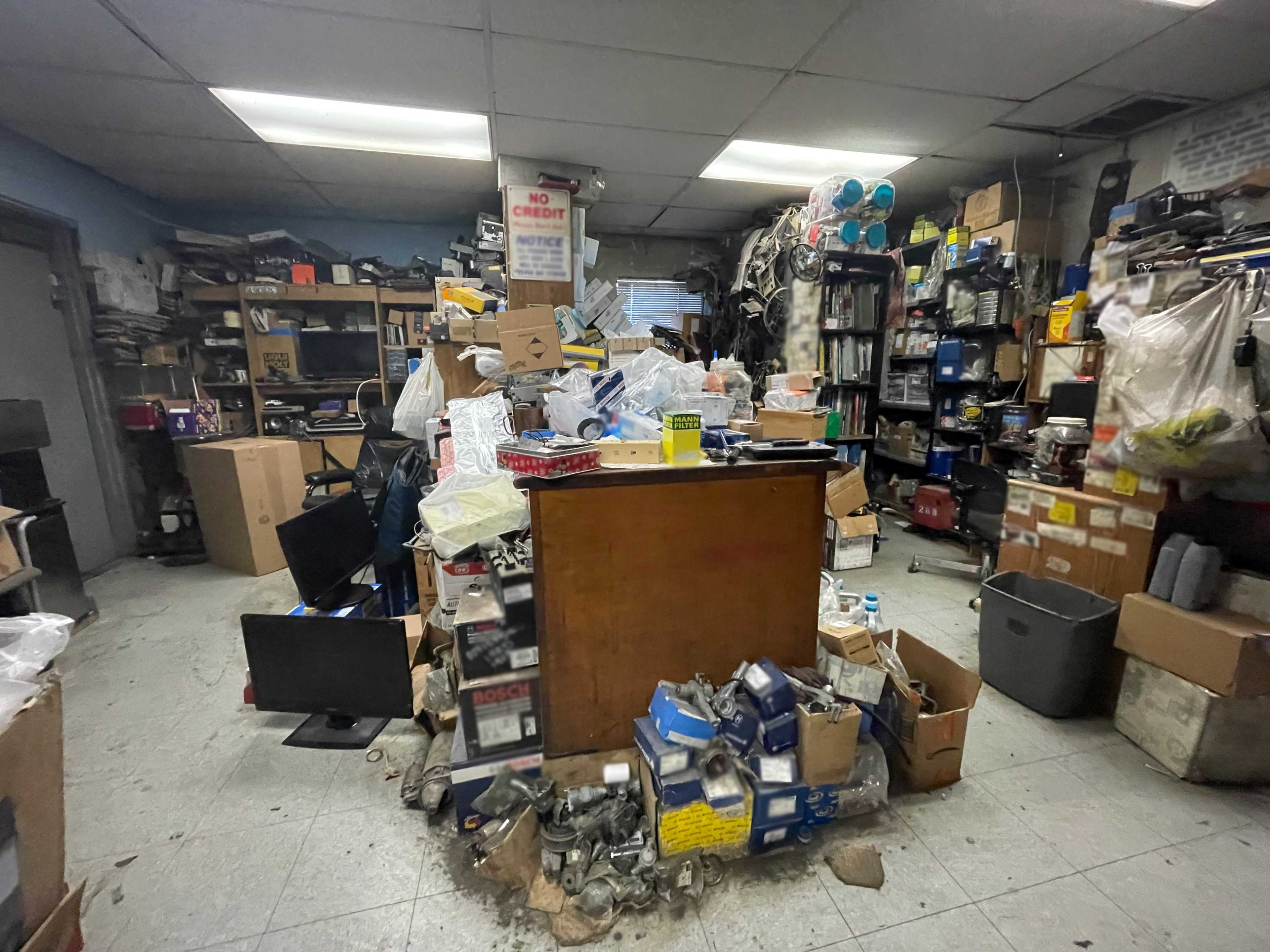 A cluttered room filled with various boxes, tools, and scattered items, with a desk at the center surrounded by stacks of materials.