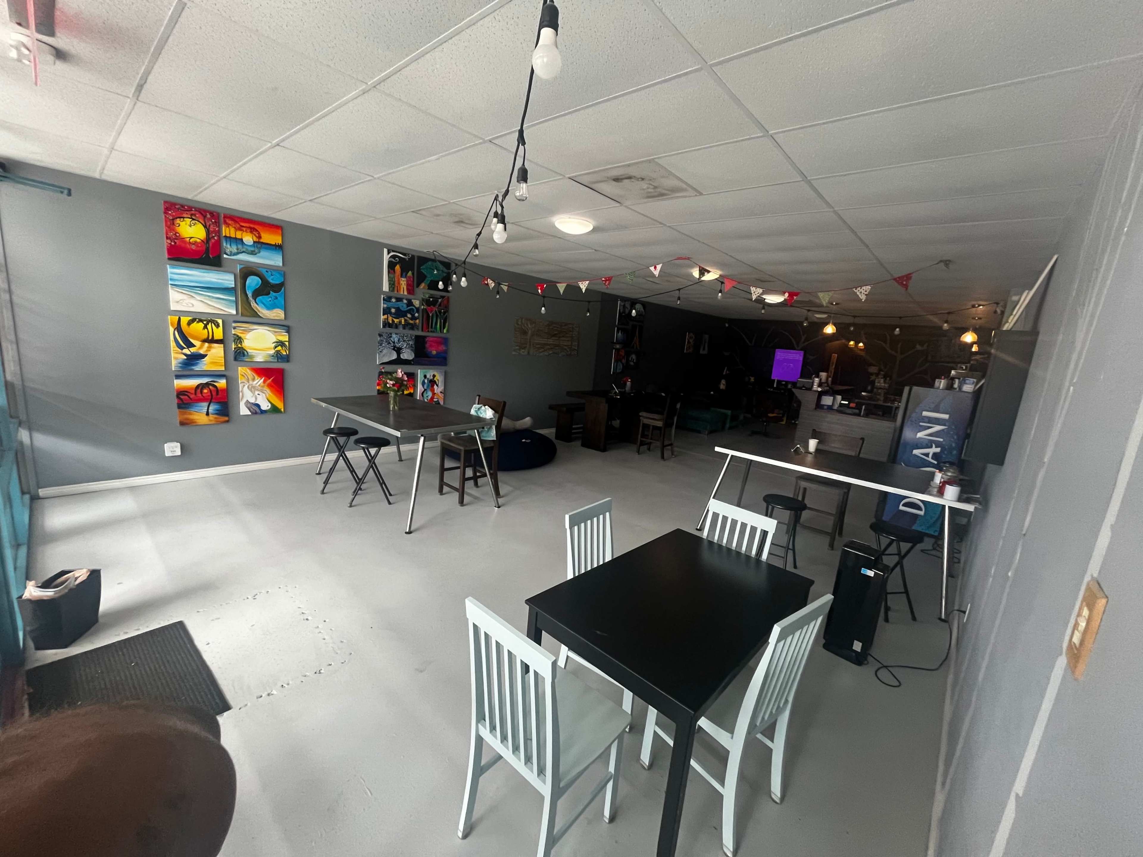 A spacious interior of a café or lounge with tables and chairs arranged neatly, colorful artwork on the walls, and string lights hanging from the ceiling.