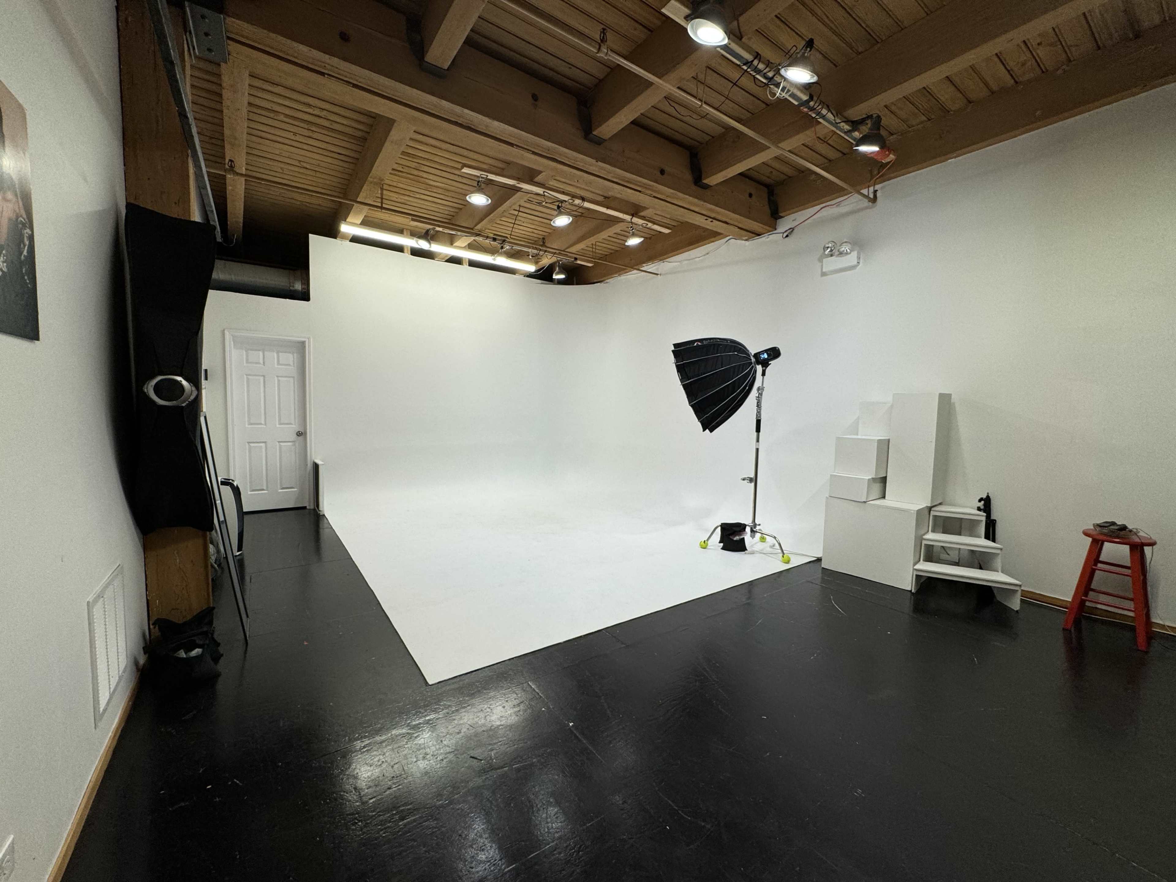 A spacious photography studio with a white backdrop, lighting equipment, and a few steps and a stool positioned in the corner.