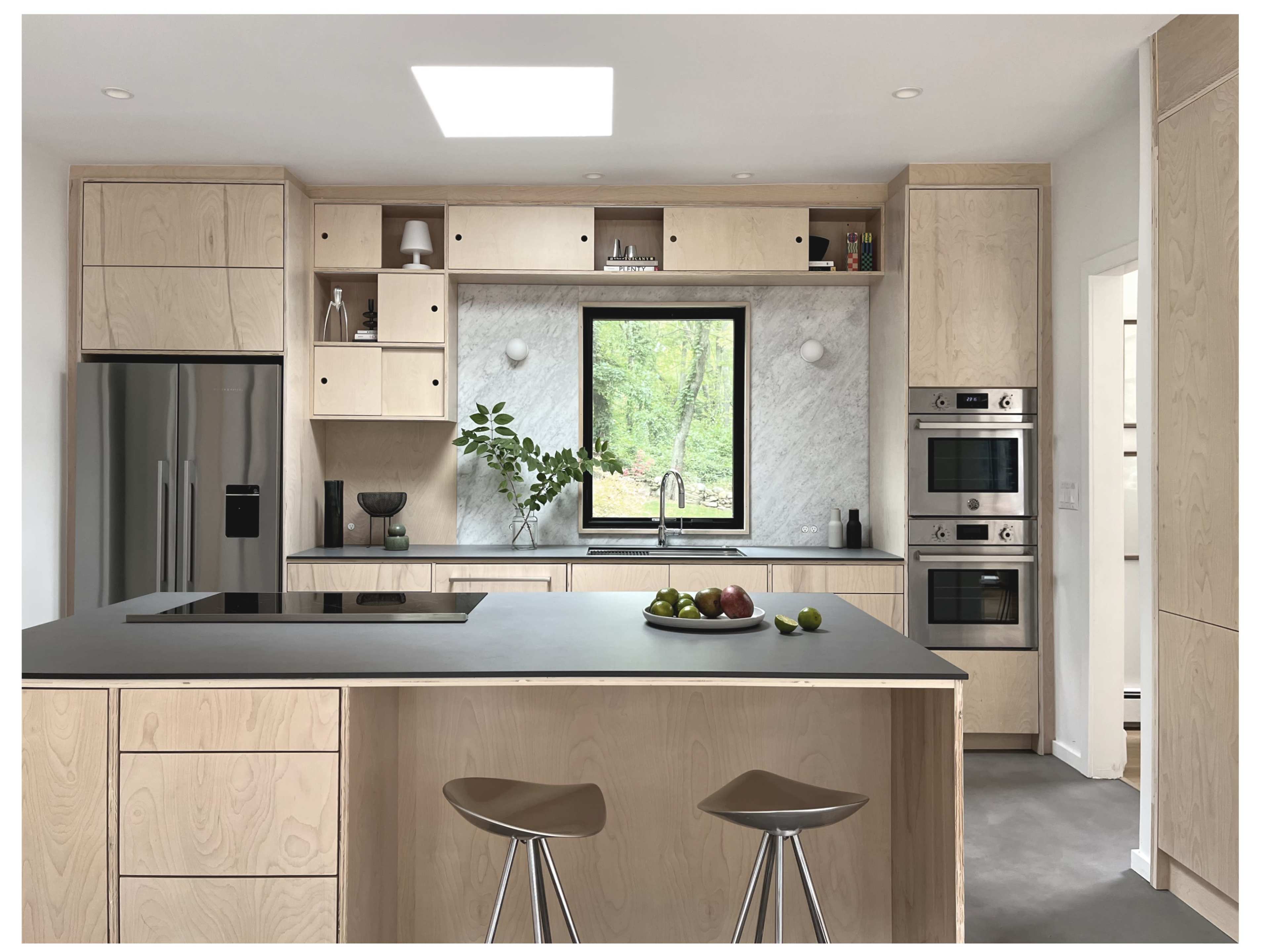 The image shows a modern kitchen featuring light wood cabinetry, a central island with a gray countertop, and large windows providing natural light and a view of greenery outside.