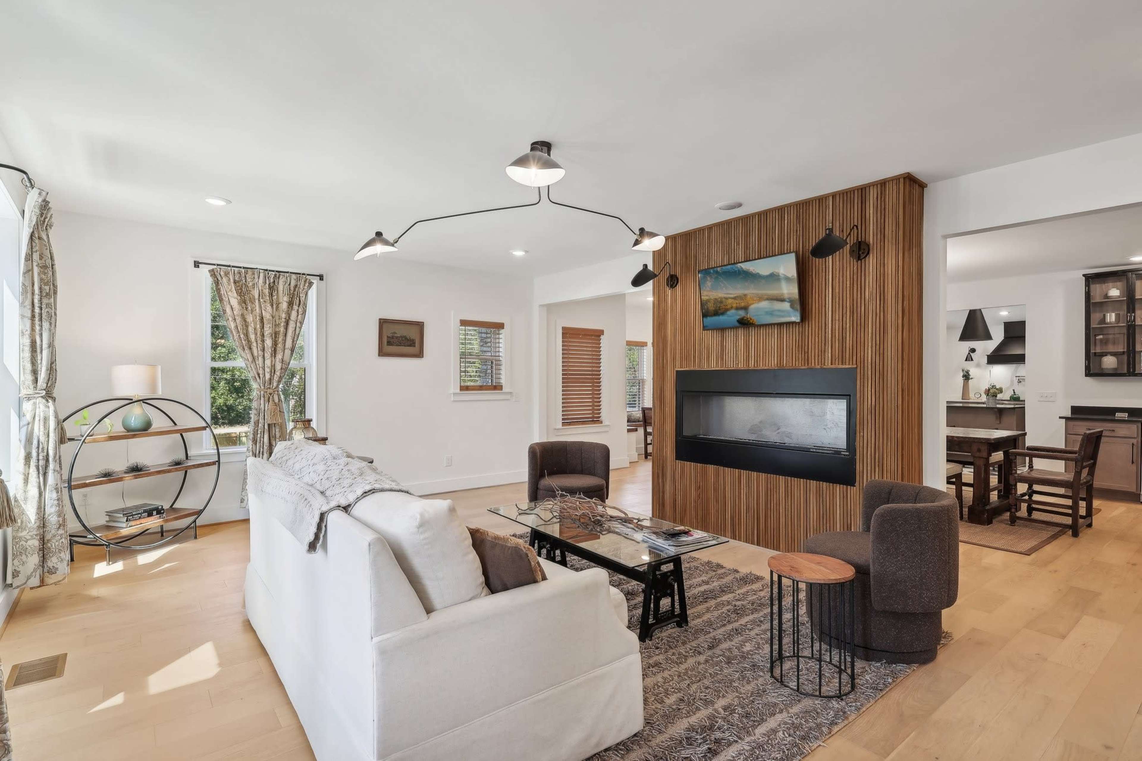 The living room features a modern design with a white sofa, a circular metal side table, and a wall-mounted television above a fireplace, set against wooden paneling.