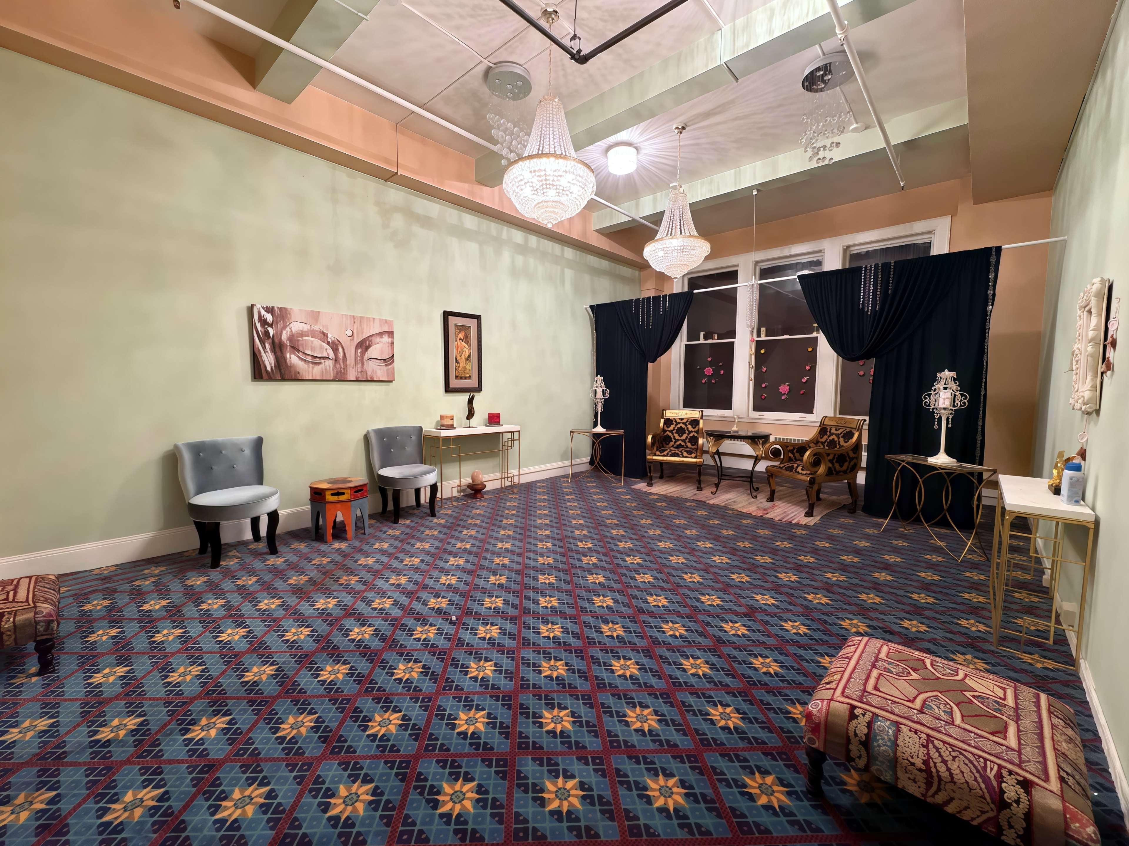 The image shows a spacious, elegantly furnished room with patterned carpet, two chandeliers, a seating area, and decorative wall art.