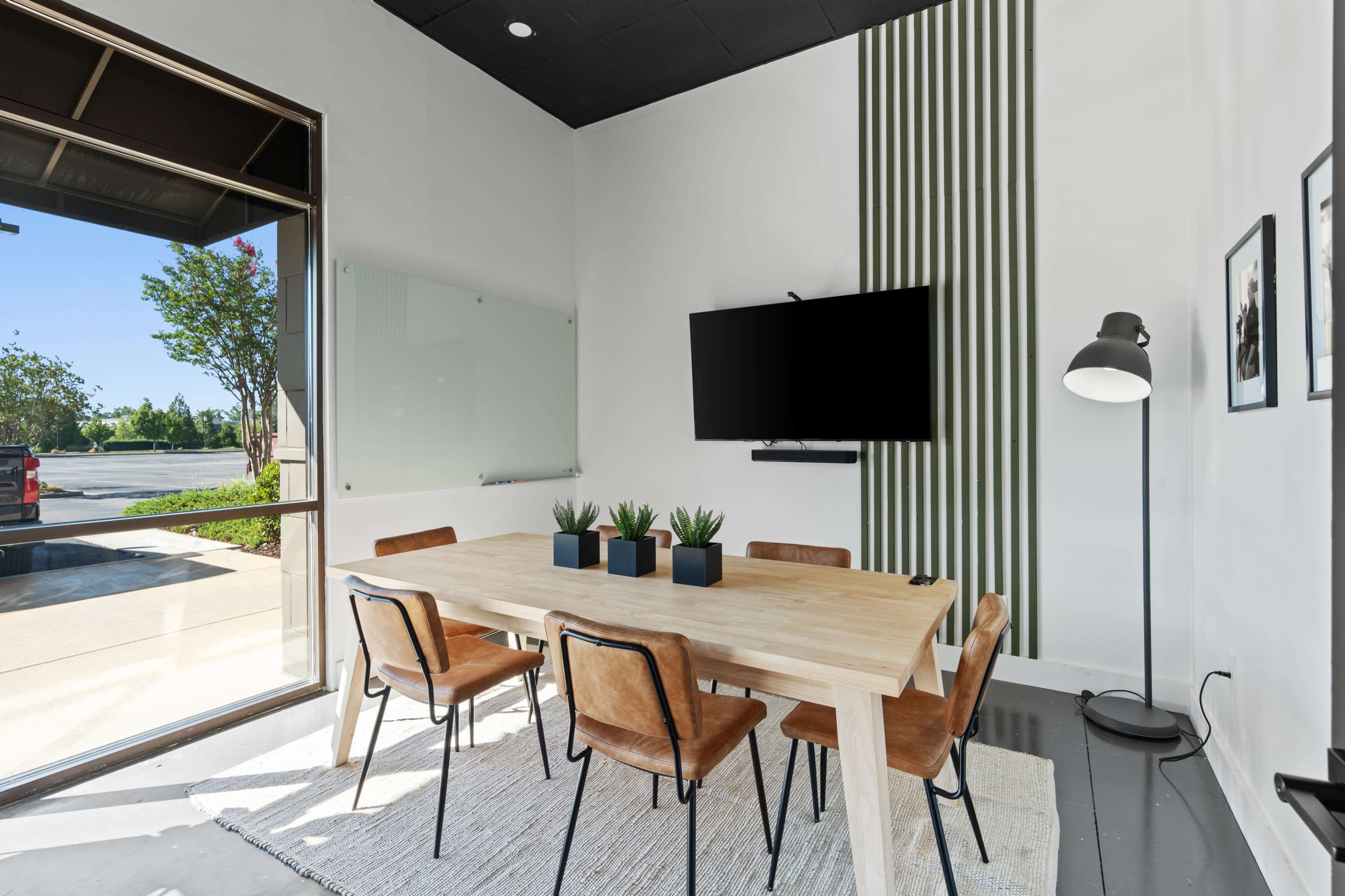 A modern conference room features a wooden table surrounded by leather chairs, with a large screen mounted on the wall and a lamp in the corner.
