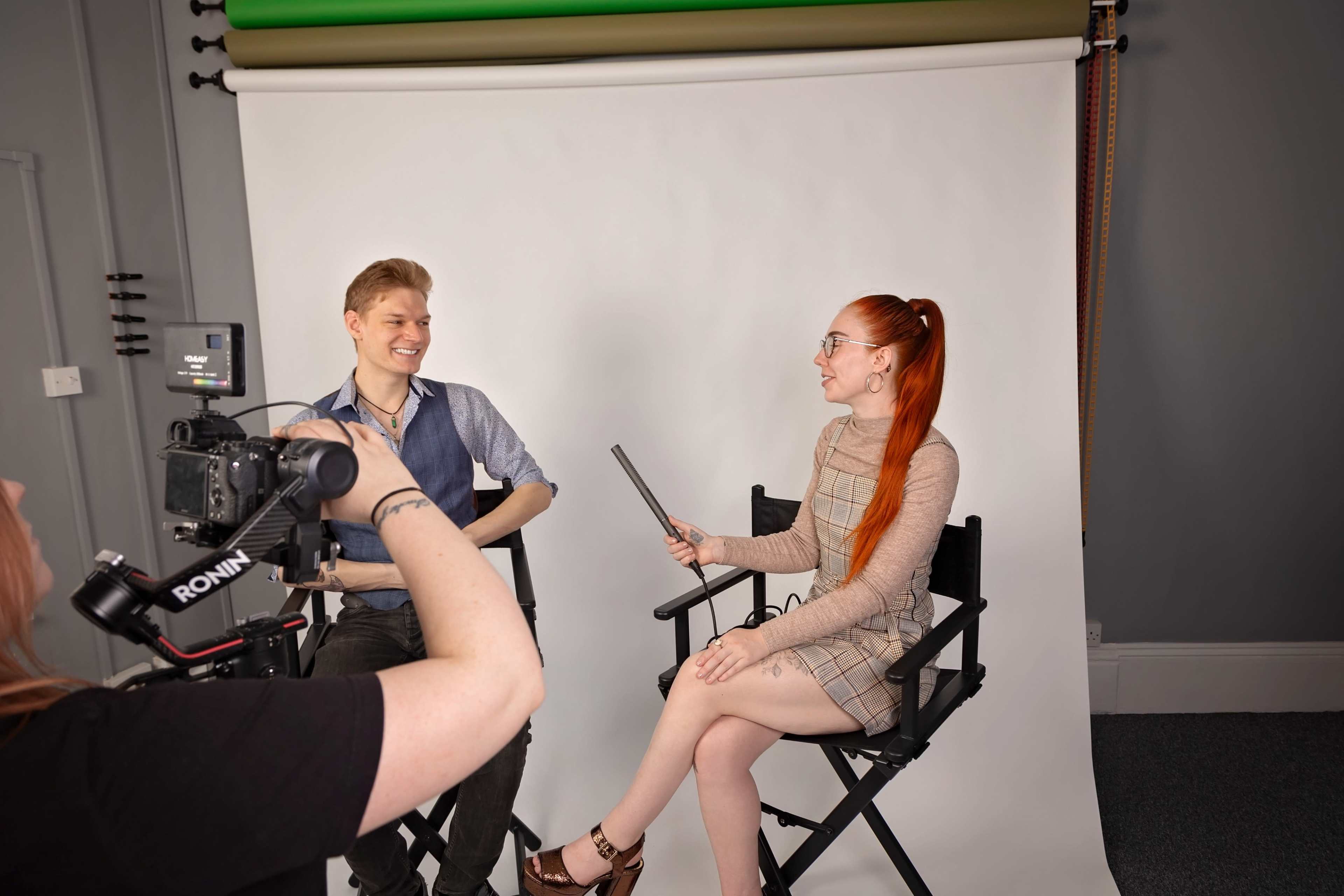 A woman interviews a man in a photography studio, with a camera on a tripod capturing the scene.