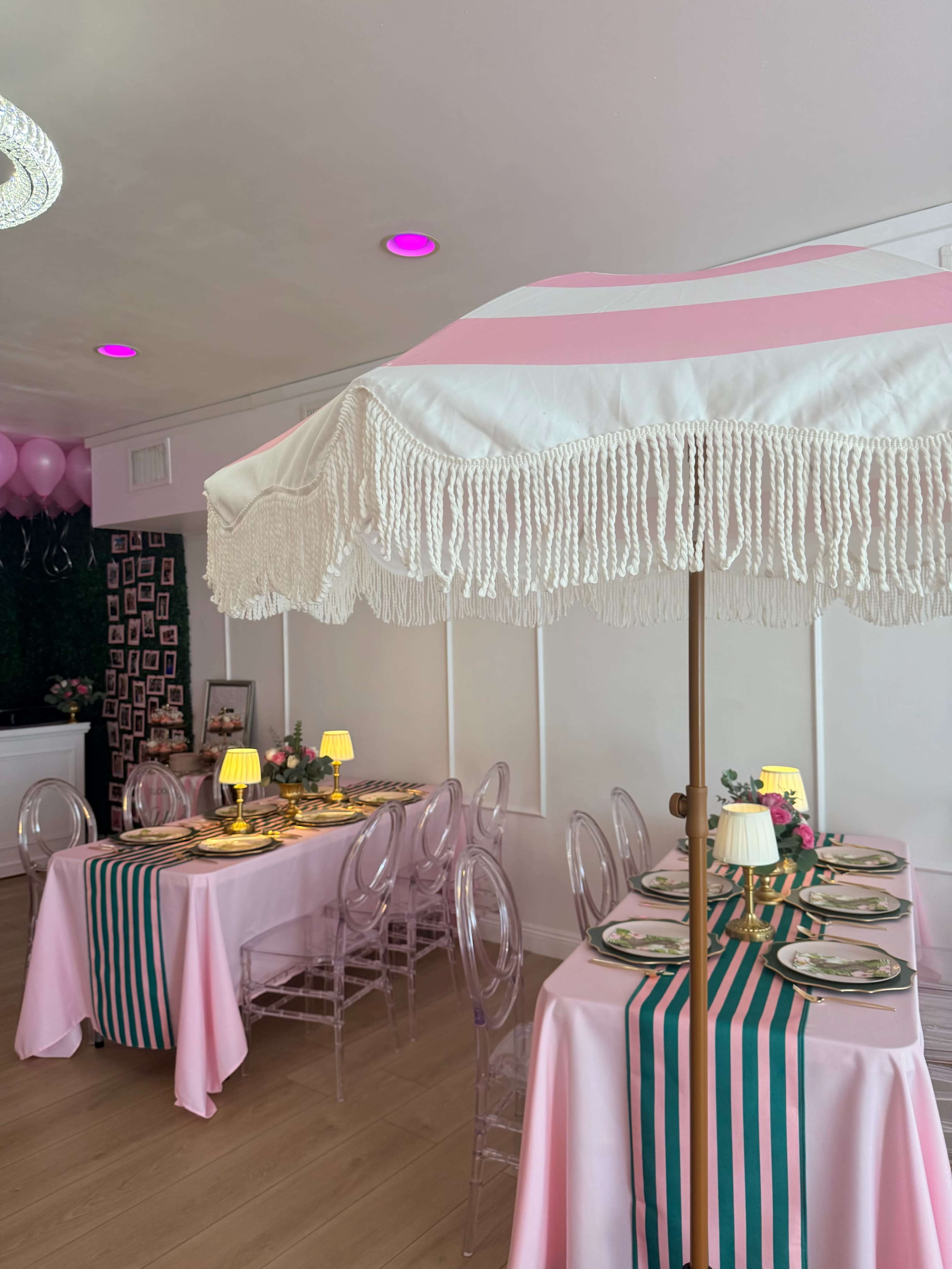 The scene depicts a brightly decorated dining area with two tables draped in pink tablecloths and striped runners, topped with umbrellas, decorative lamps, and floral centerpieces.