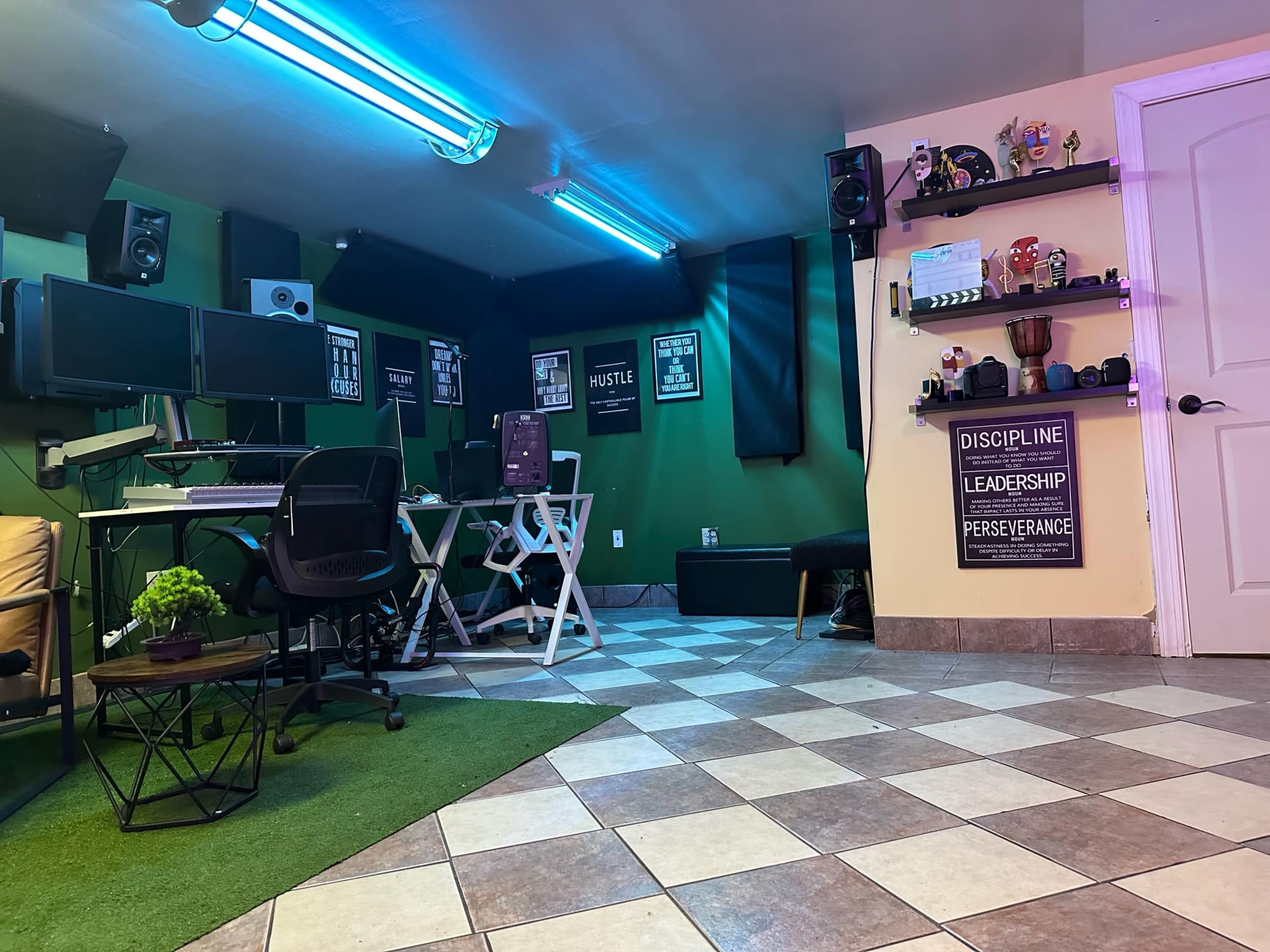 A home office setup with dual computer monitors, speakers, and motivational posters on a green-painted wall, alongside a tiled floor with a small area rug.