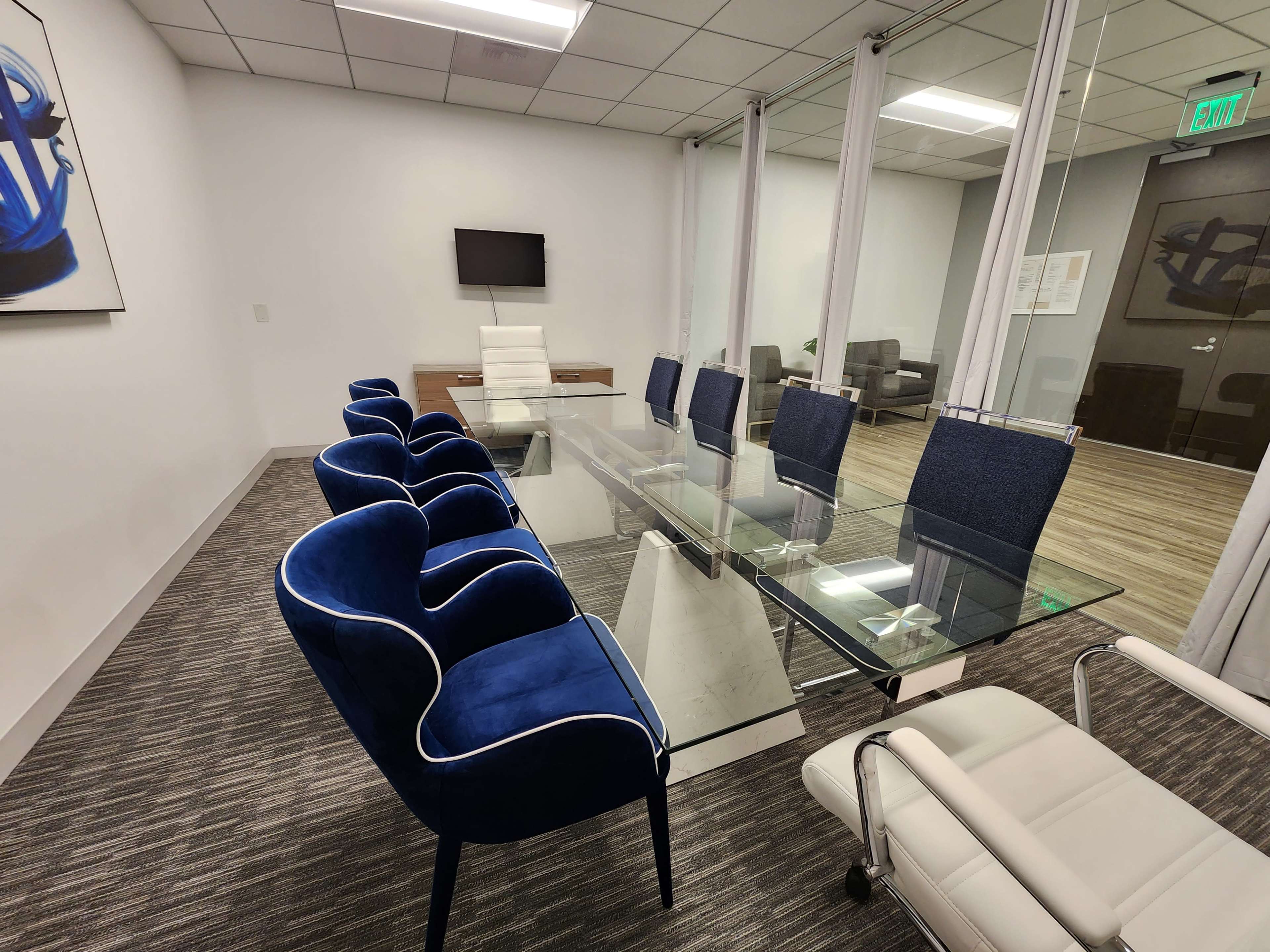 A conference room features a glass table surrounded by blue upholstered chairs and a white leather office chair, with a wall-mounted television and modern decor.