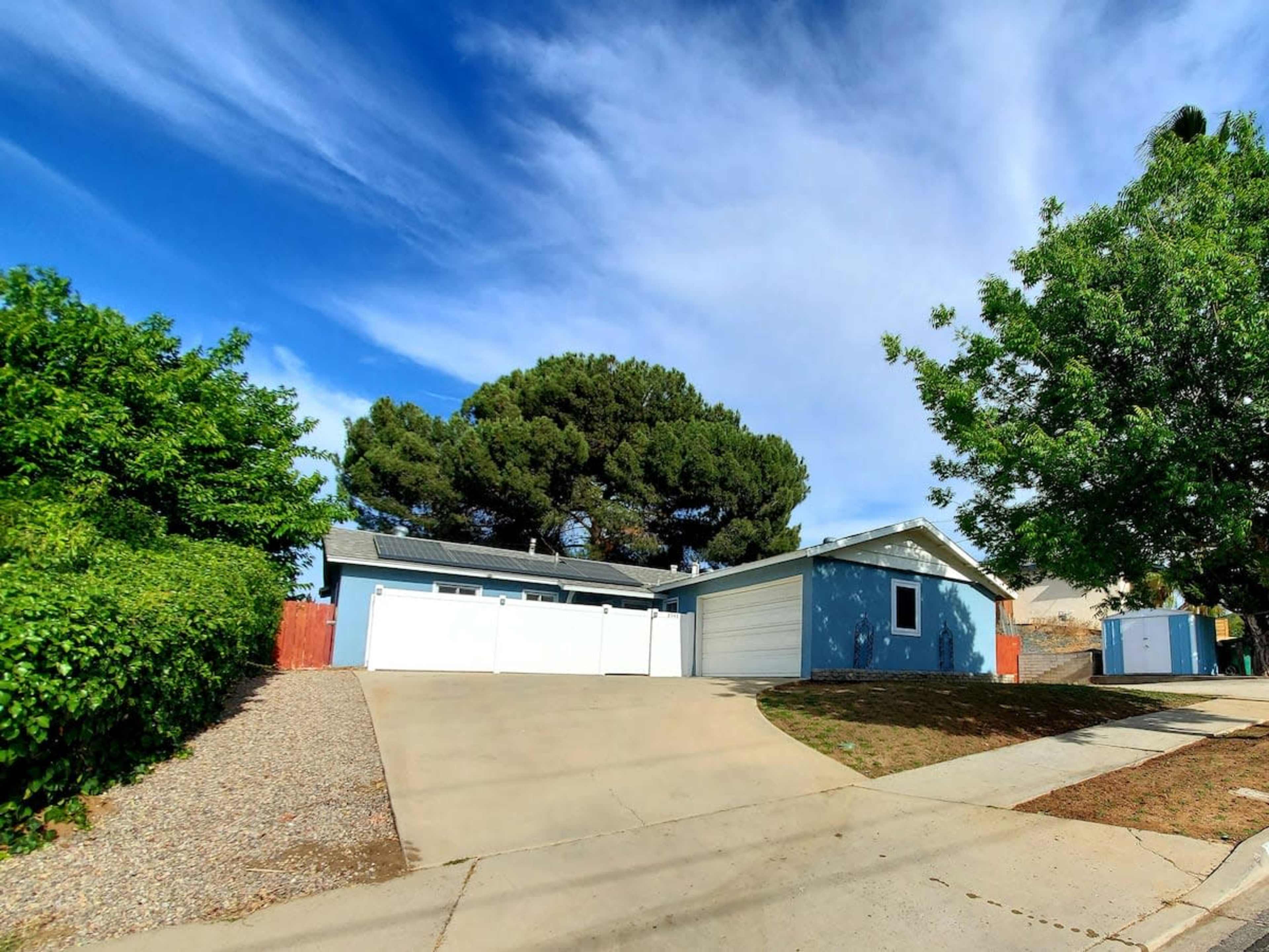 A blue house with a driveway and white garage door is surrounded by green shrubs and tall trees.