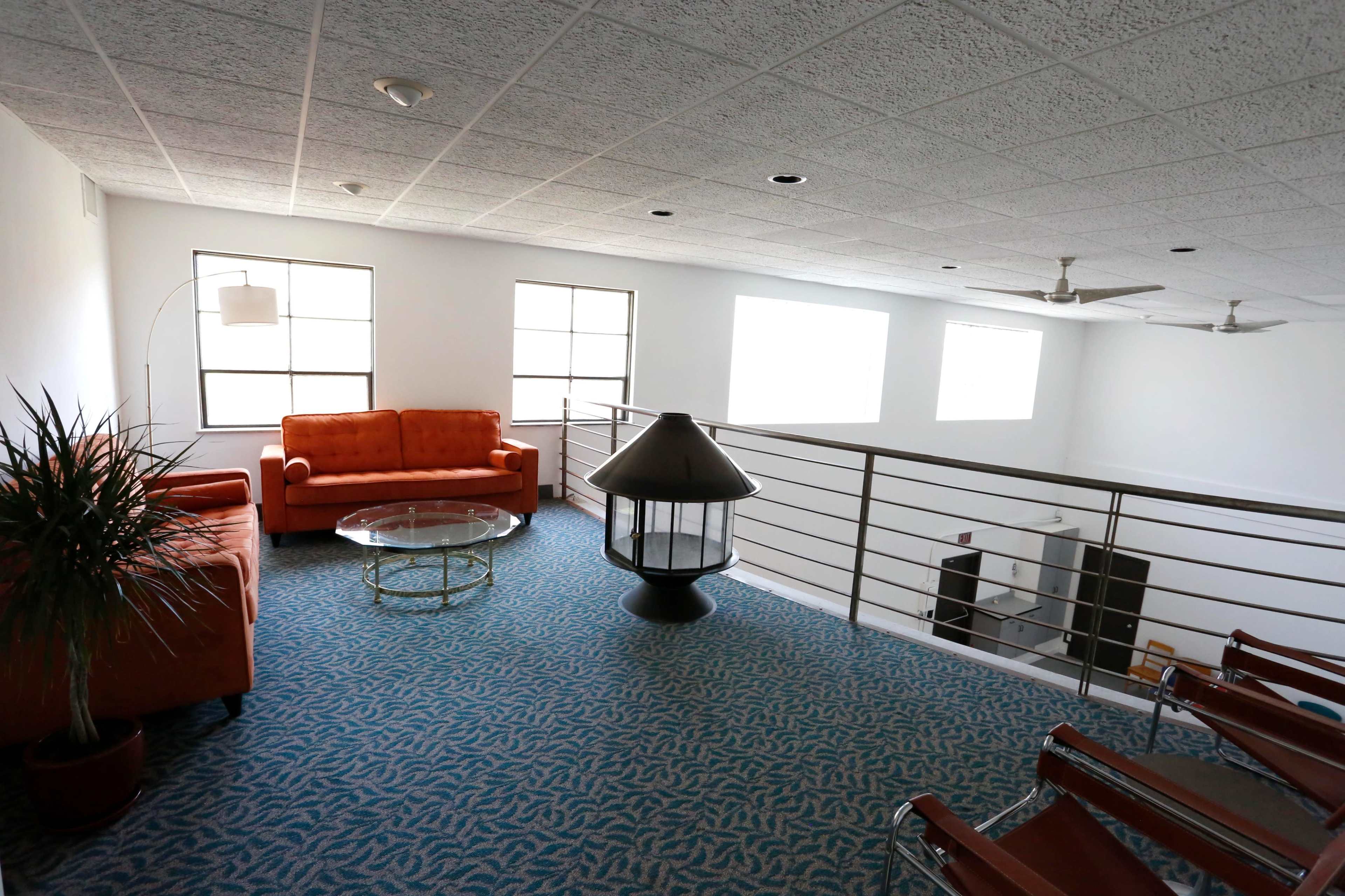 A bright, spacious loft area features a small seating arrangement with orange furniture, a glass coffee table, and a unique lamp next to a railing overlooking the lower level.