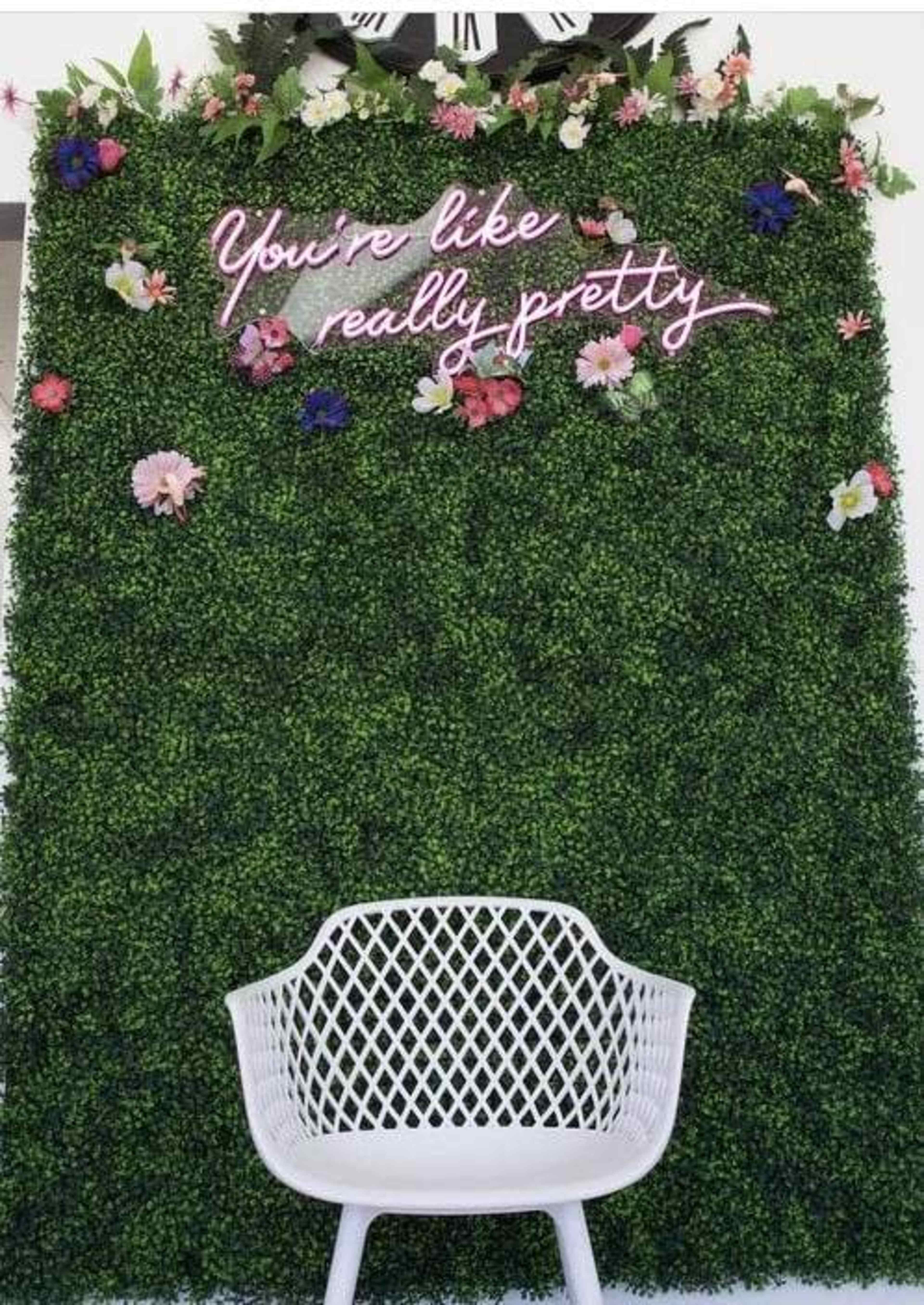 A white chair is positioned in front of a green foliage wall adorned with flowers and a neon sign that reads "You're like really pretty."