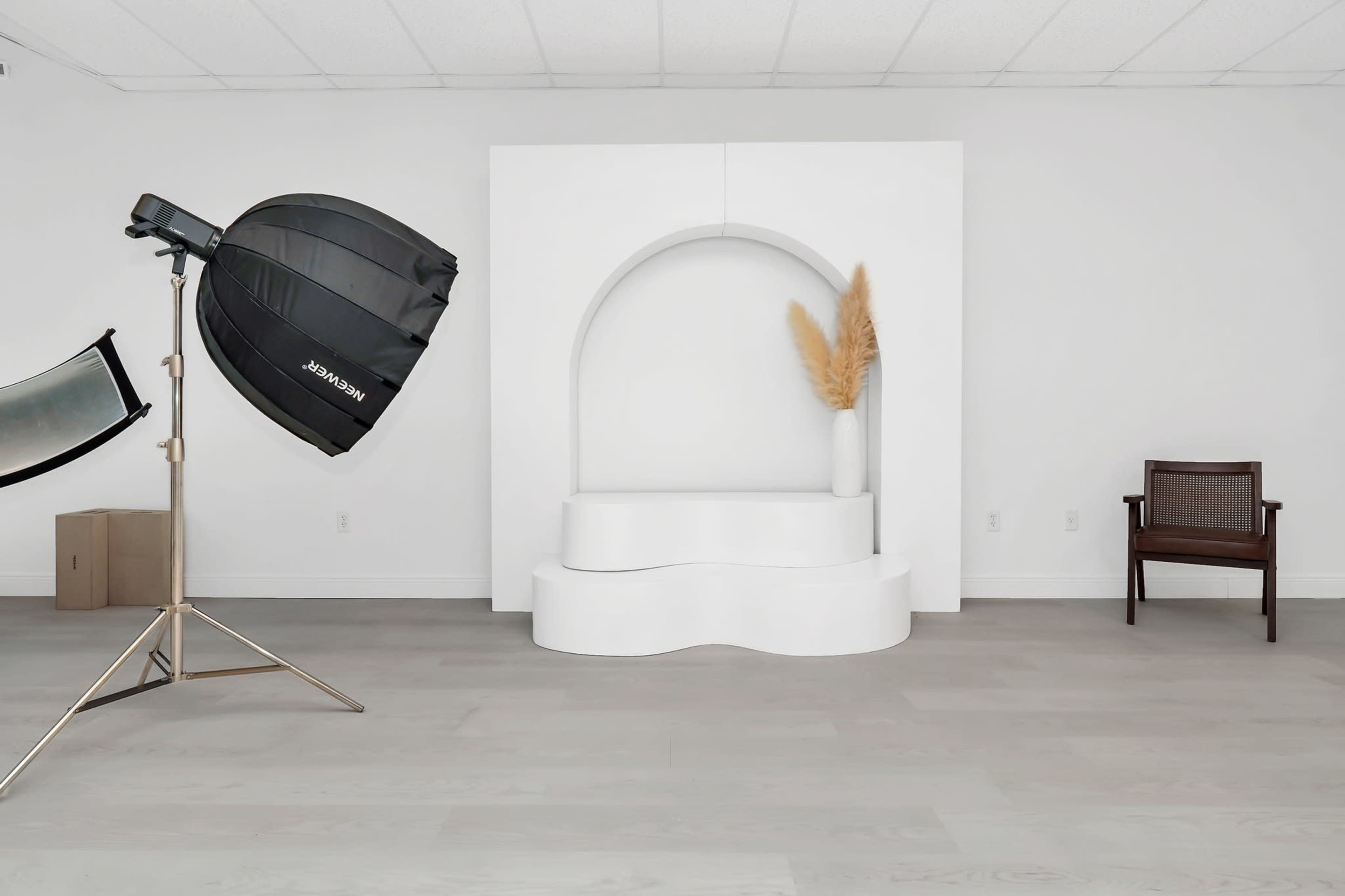 The image shows a minimalist studio setup featuring a large black softbox light, a curved white display stand with a plant, and a wooden chair against a plain white wall.