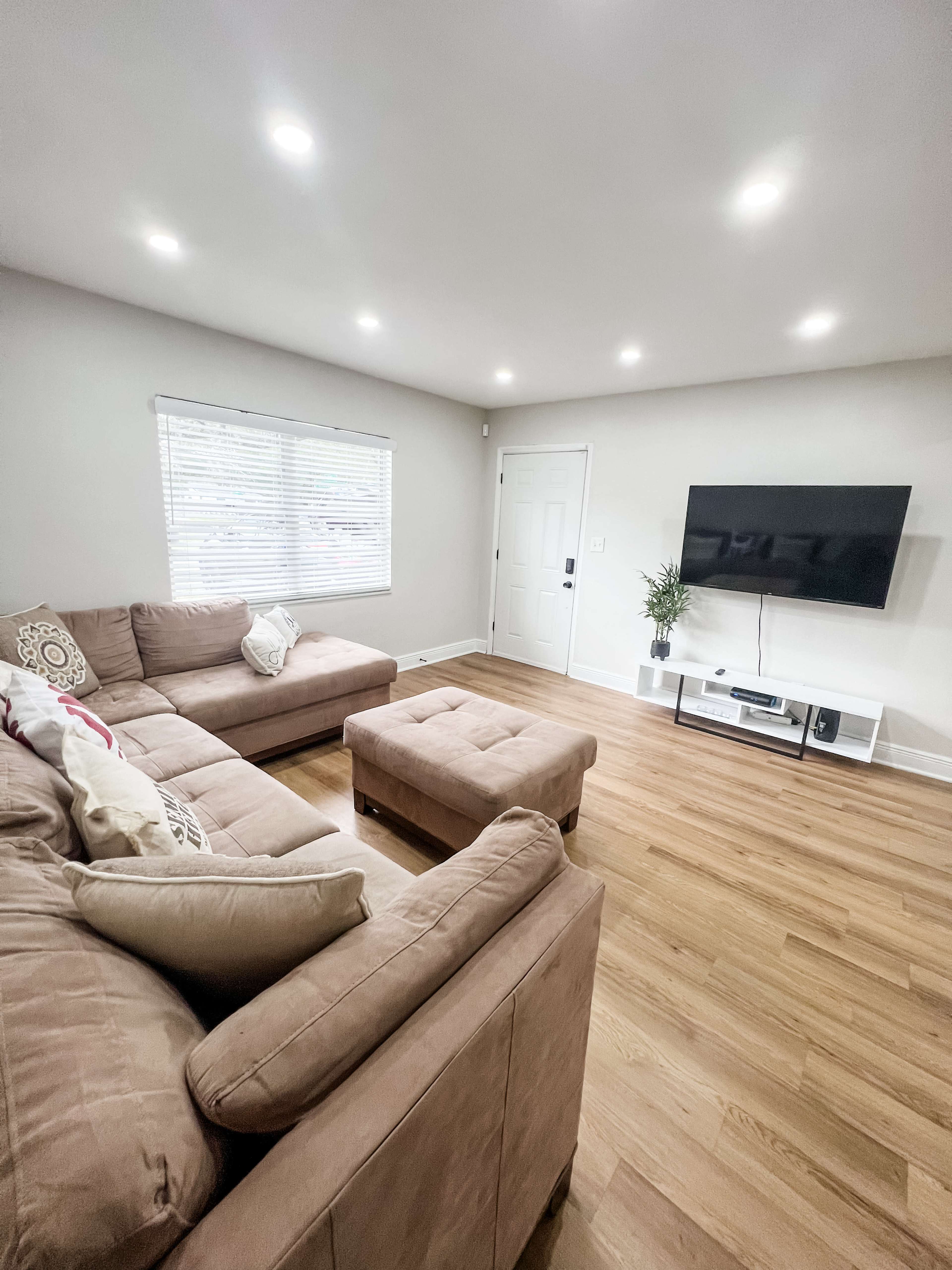A living room with a large sectional sofa, a coffee table, a television mounted on the wall, and a window with blinds.
