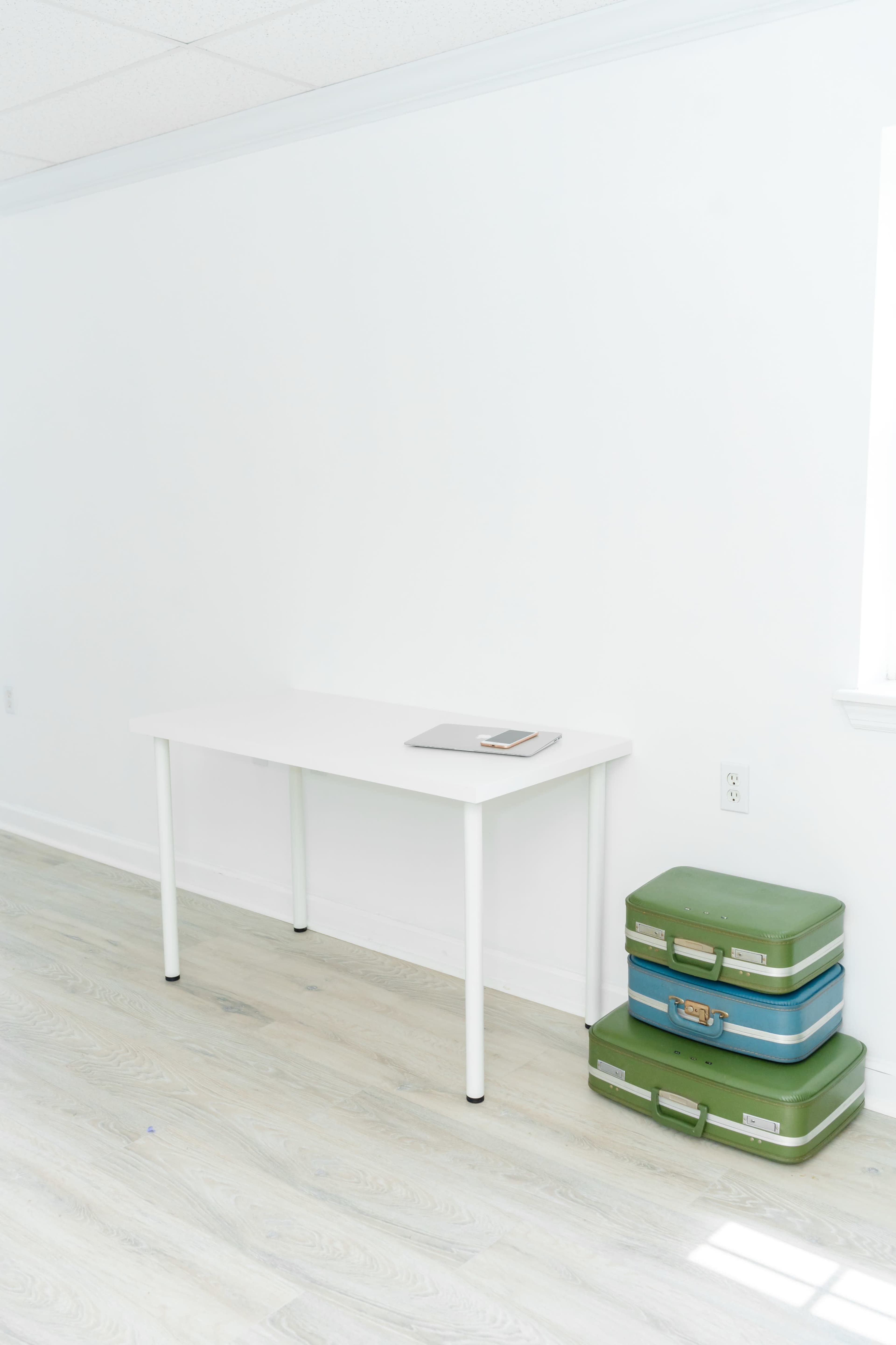 A white table with a laptop sits against a blank wall, next to a stack of green suitcases.