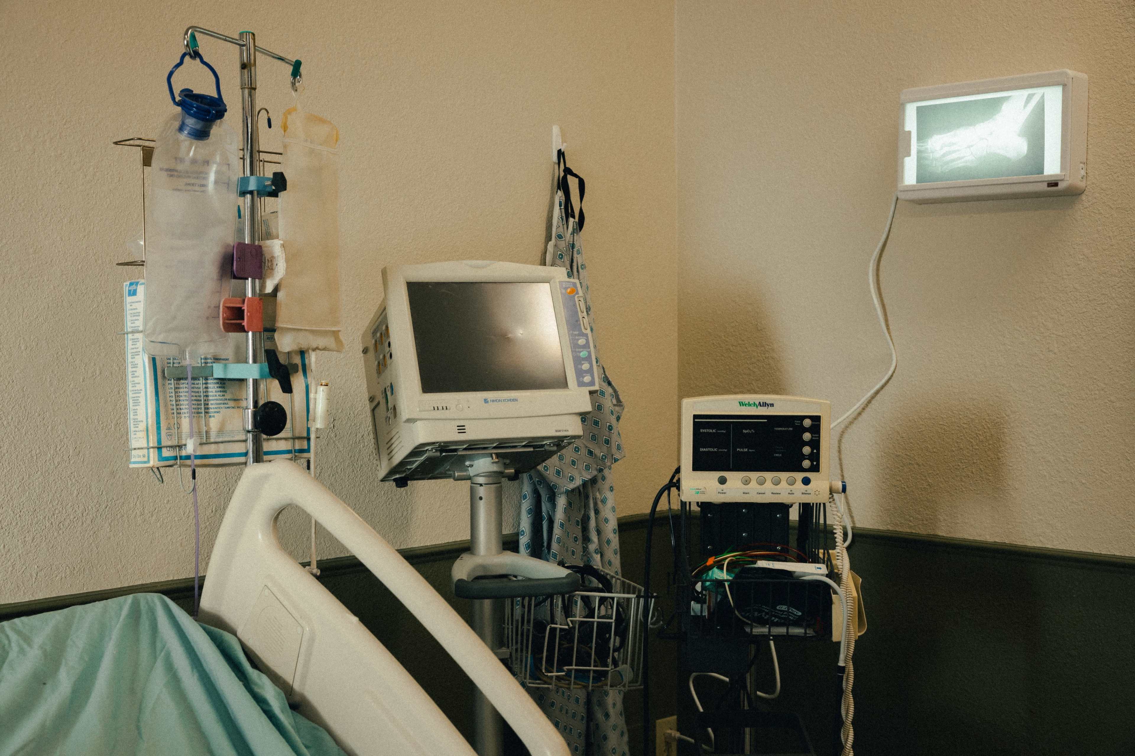 The image shows a hospital room with a medical bed, monitoring equipment, and an X-ray displayed on the wall.