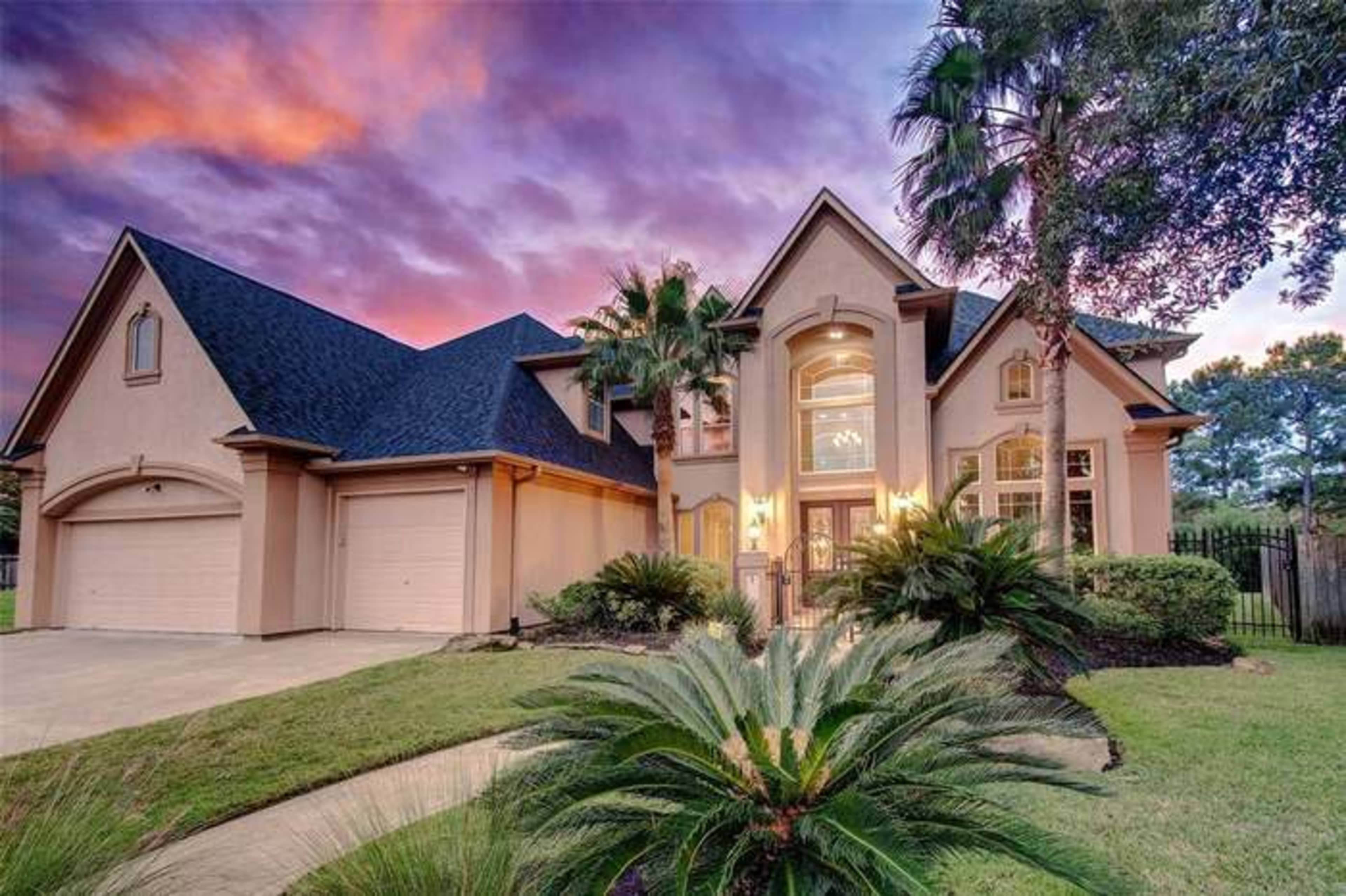 A large, modern two-story house with a landscaped yard and palm trees stands under a vibrant sunset sky.