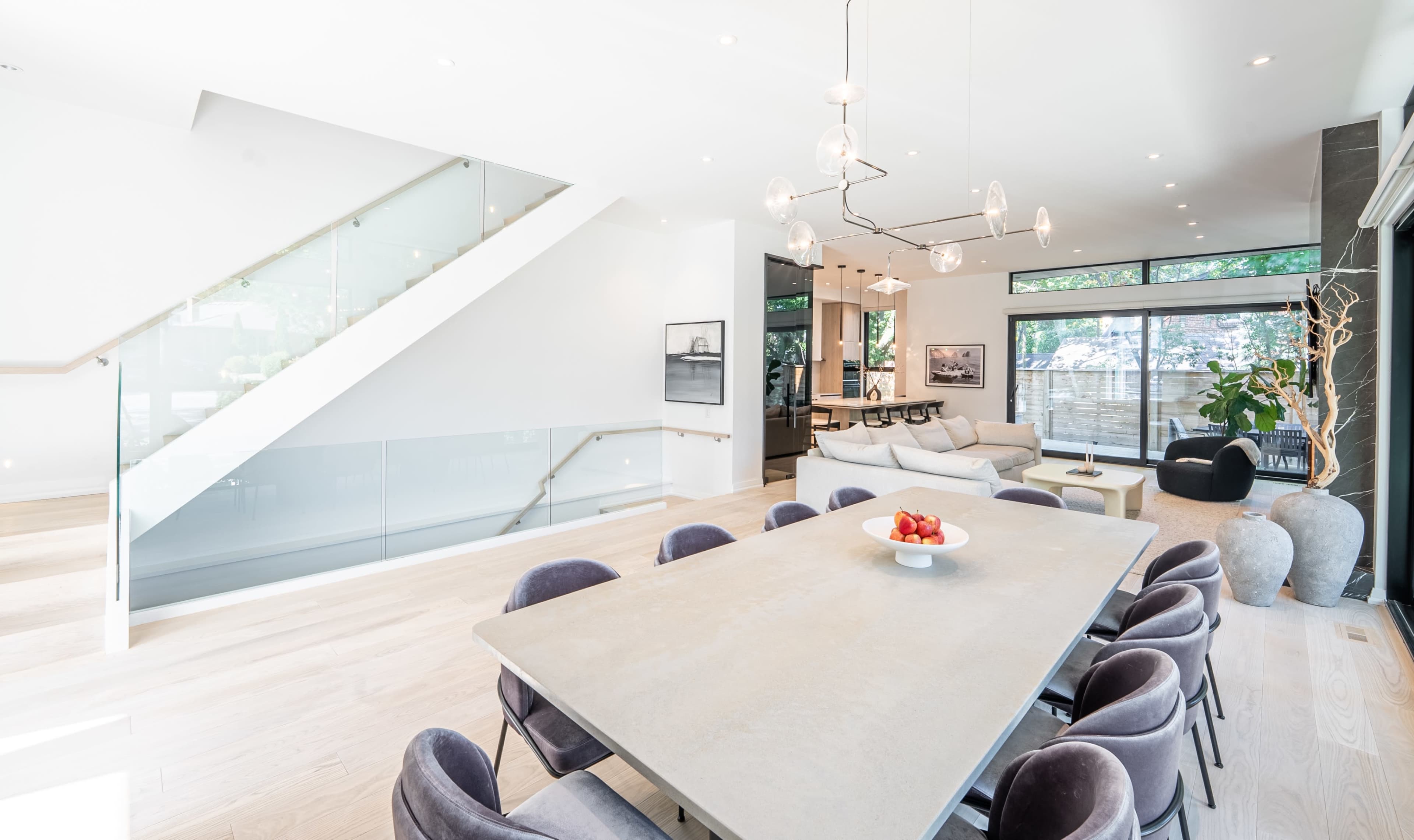 The image shows a modern dining area with a large table surrounded by upholstered chairs, adjacent to a staircase with glass railings and a living space featuring large windows.
