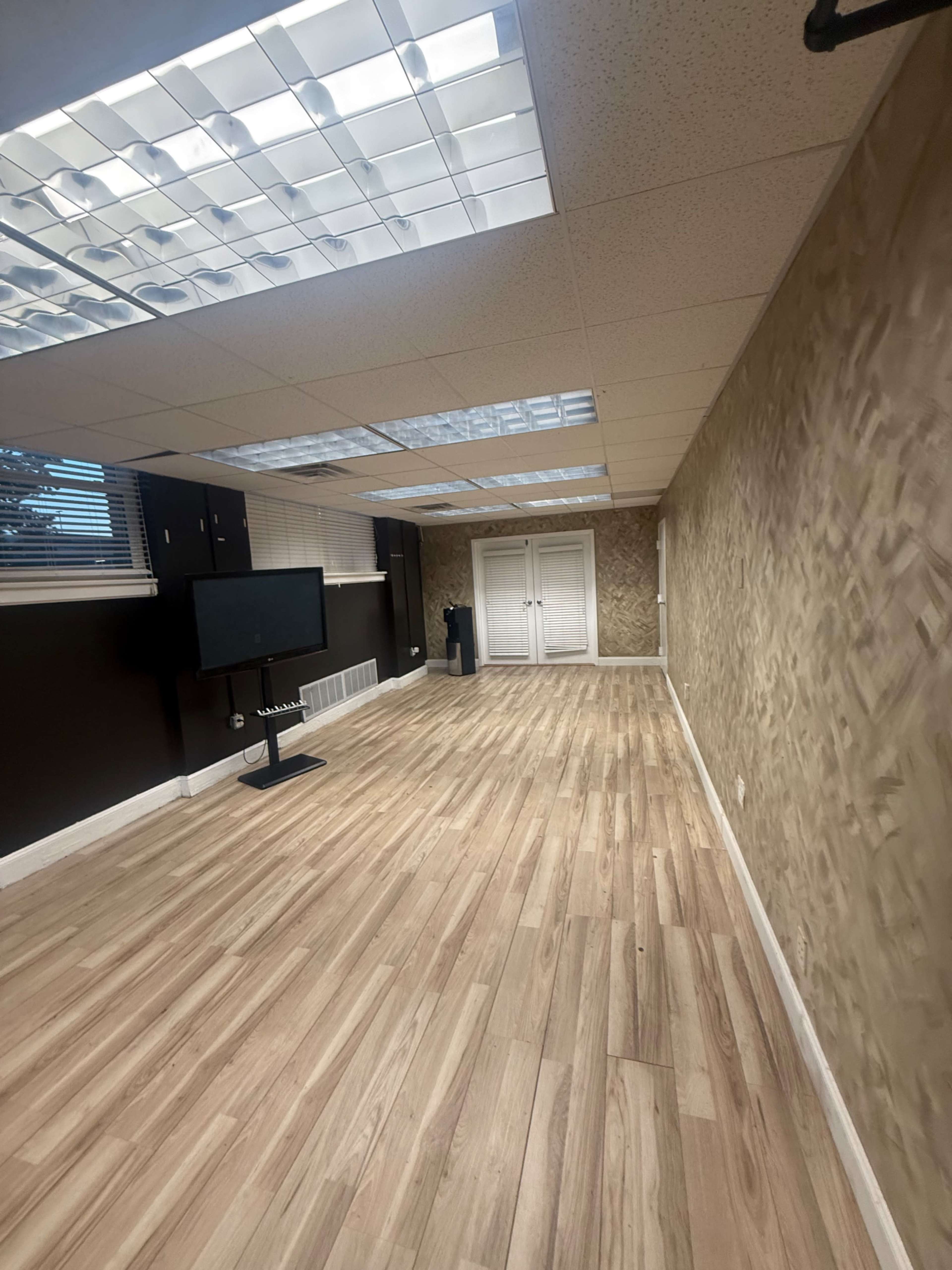 The image shows a spacious, empty room with wooden flooring, a large window, and a television mounted on a stand.
