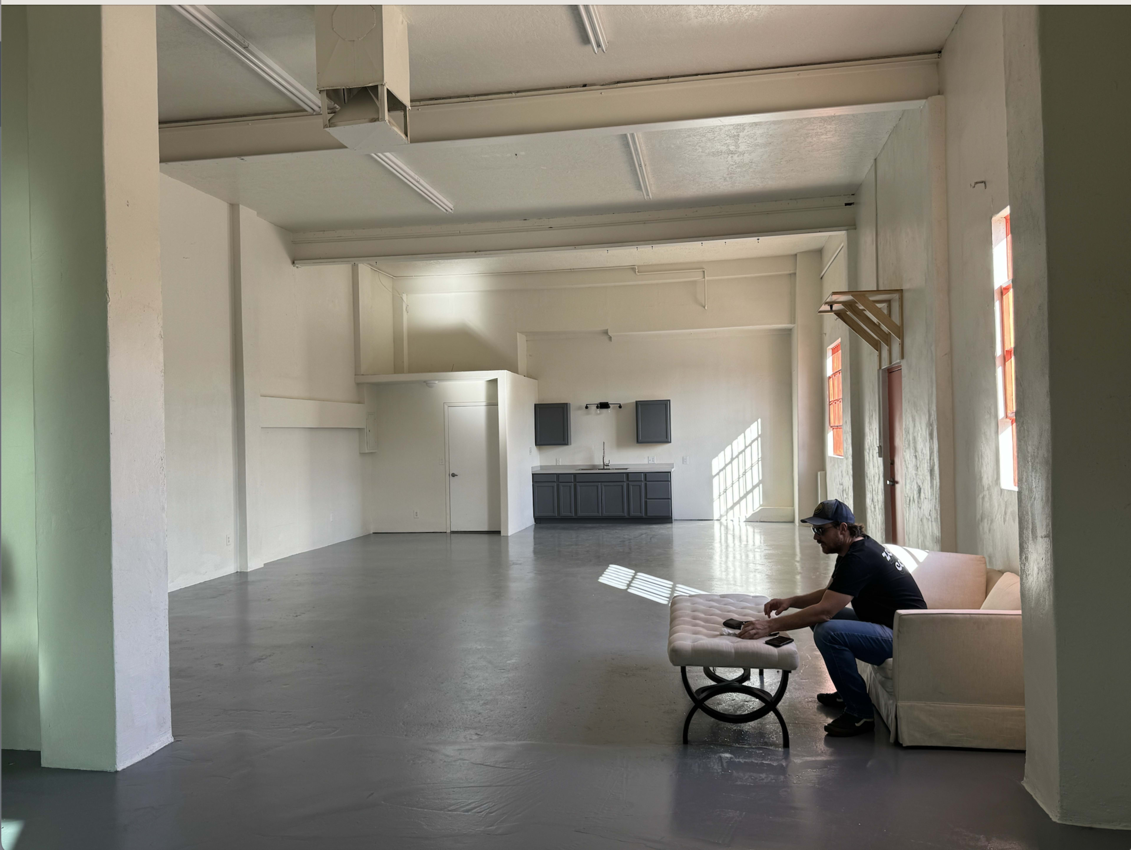 A man sits on a couch in a spacious, empty room with white walls and large windows, featuring a kitchenette in the background.