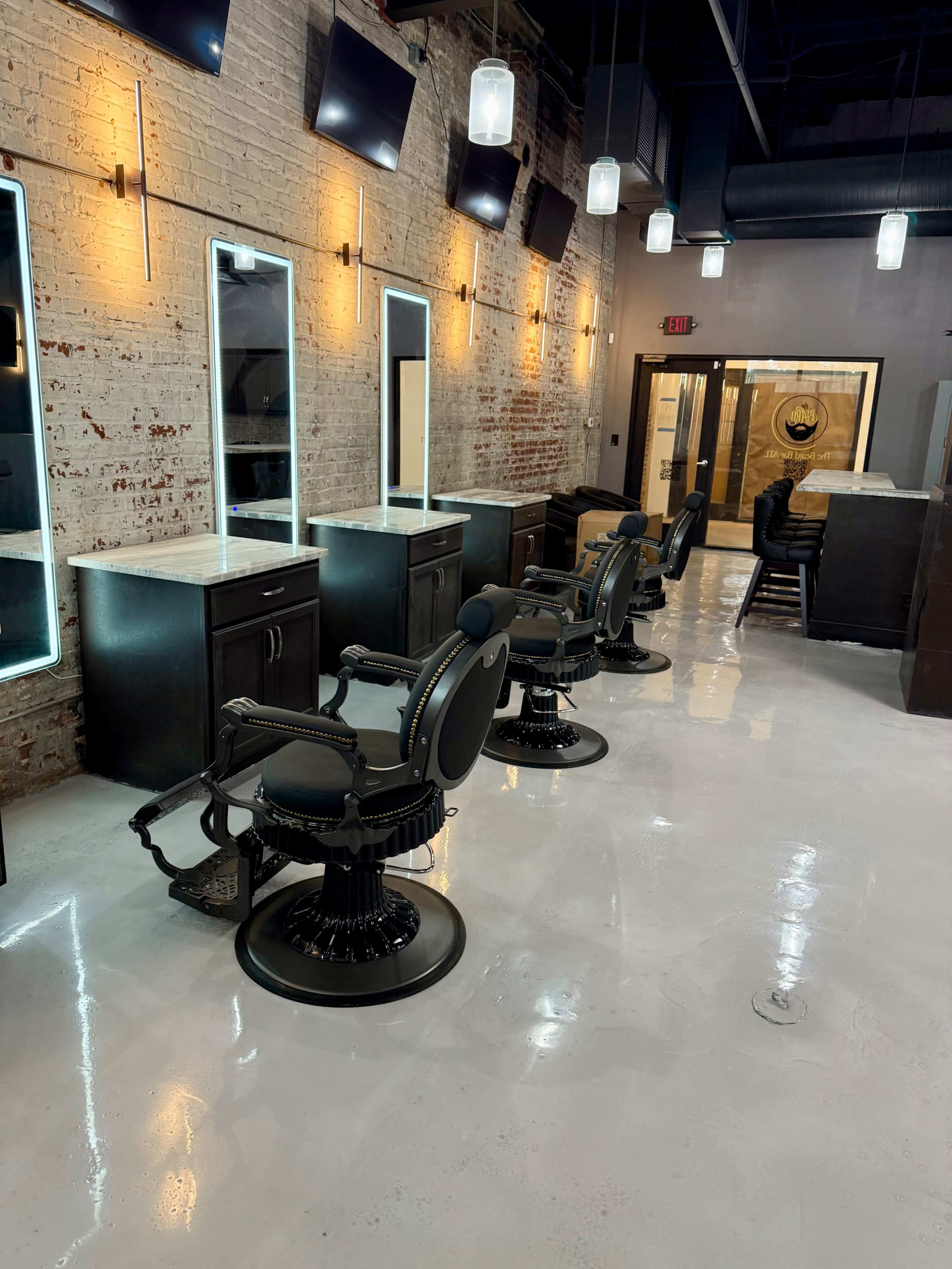 The image shows a modern barber shop with several styling chairs lined up in front of illuminated mirrors against a brick wall.