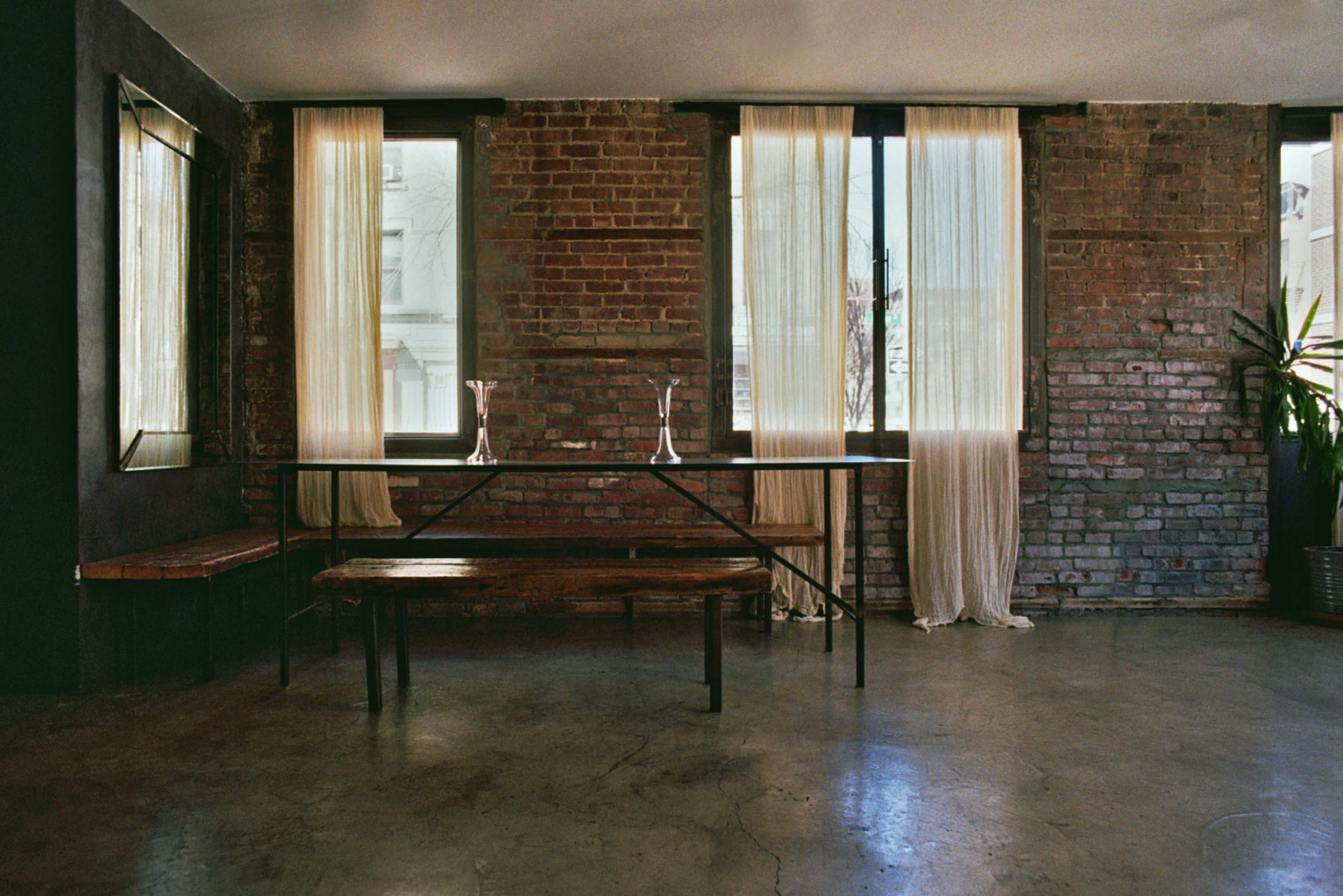 The image shows a minimalistic interior space featuring a table and bench made of wood, with large windows draped in light curtains, exposed brick walls, and a potted plant.