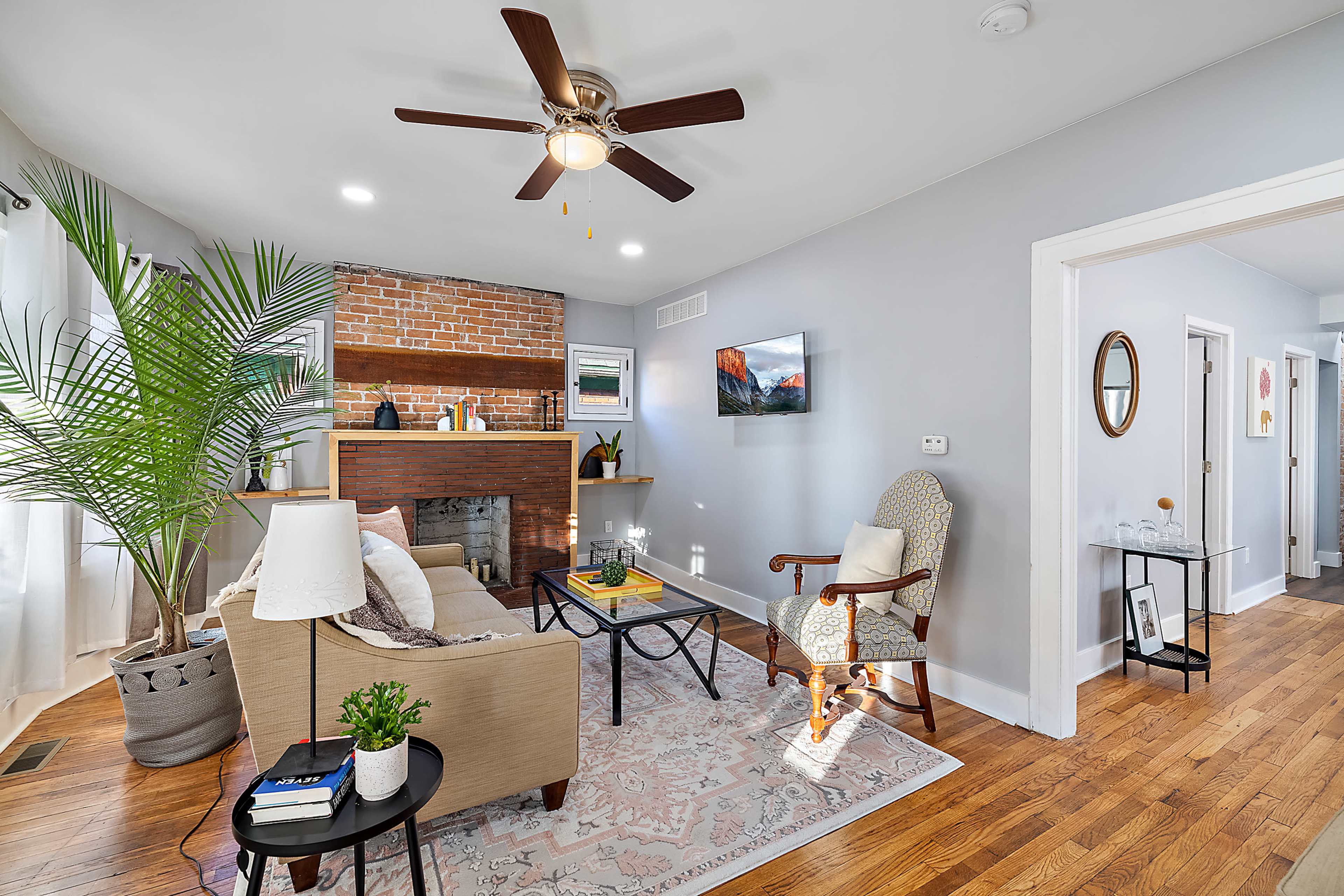 The image shows a cozy living room with a ceiling fan, a brick fireplace, and a mix of modern and traditional furnishings.
