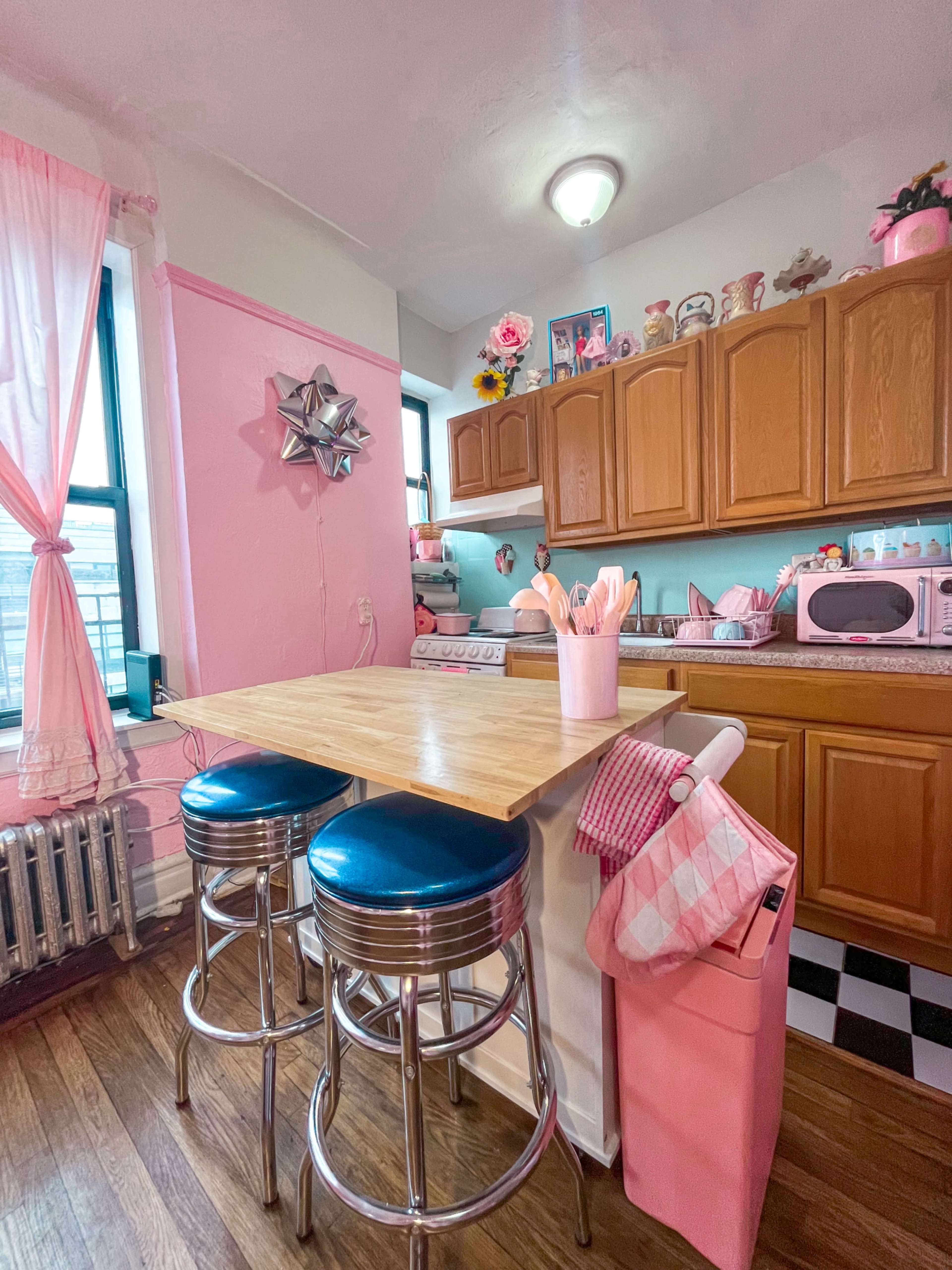 The kitchen features light pink walls, wooden cabinets, and a small table with two chrome stools.