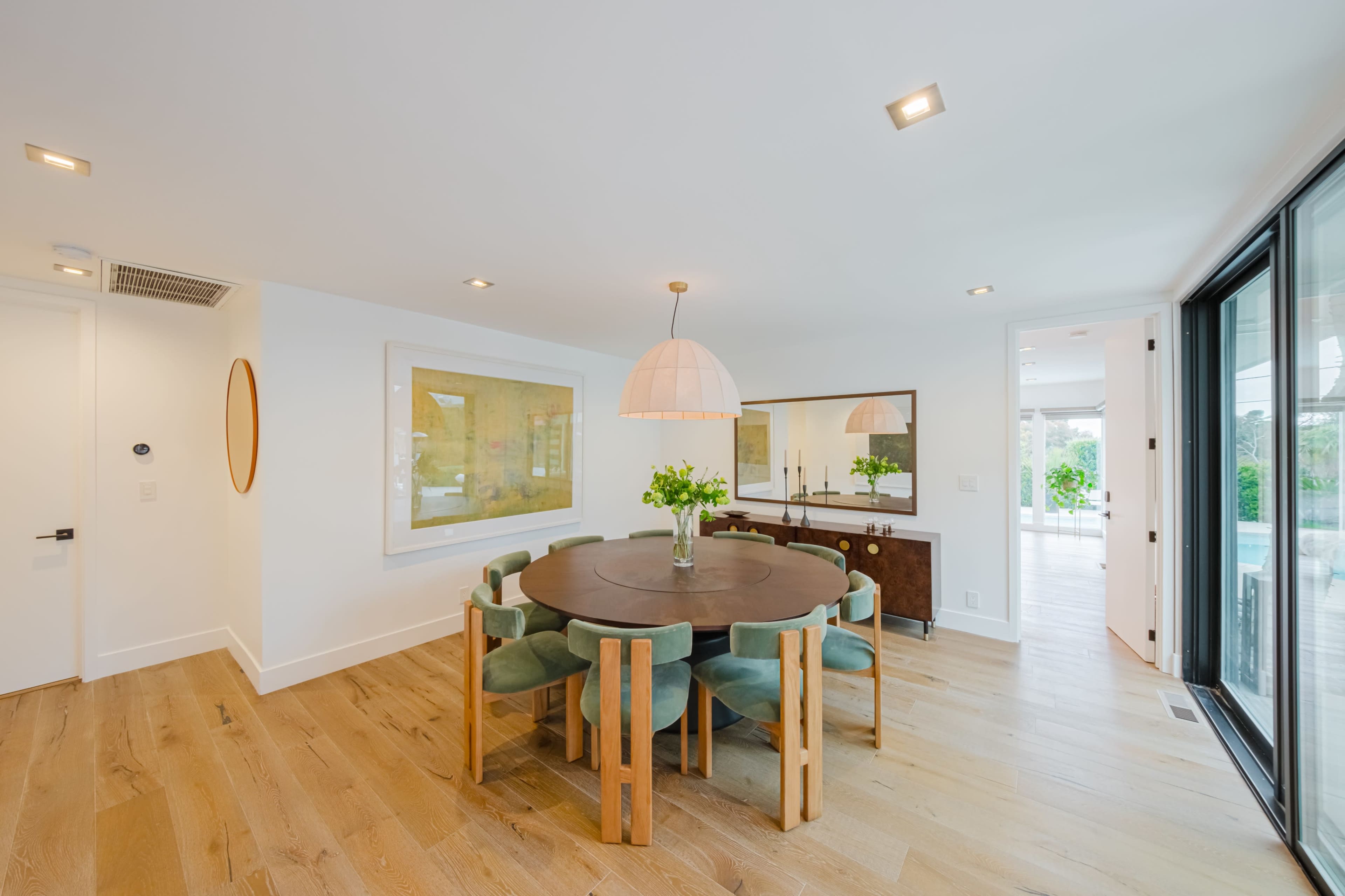 The image shows a modern dining area featuring a round wooden table surrounded by chairs, a large artwork on the wall, and glass doors leading to an outdoor space.