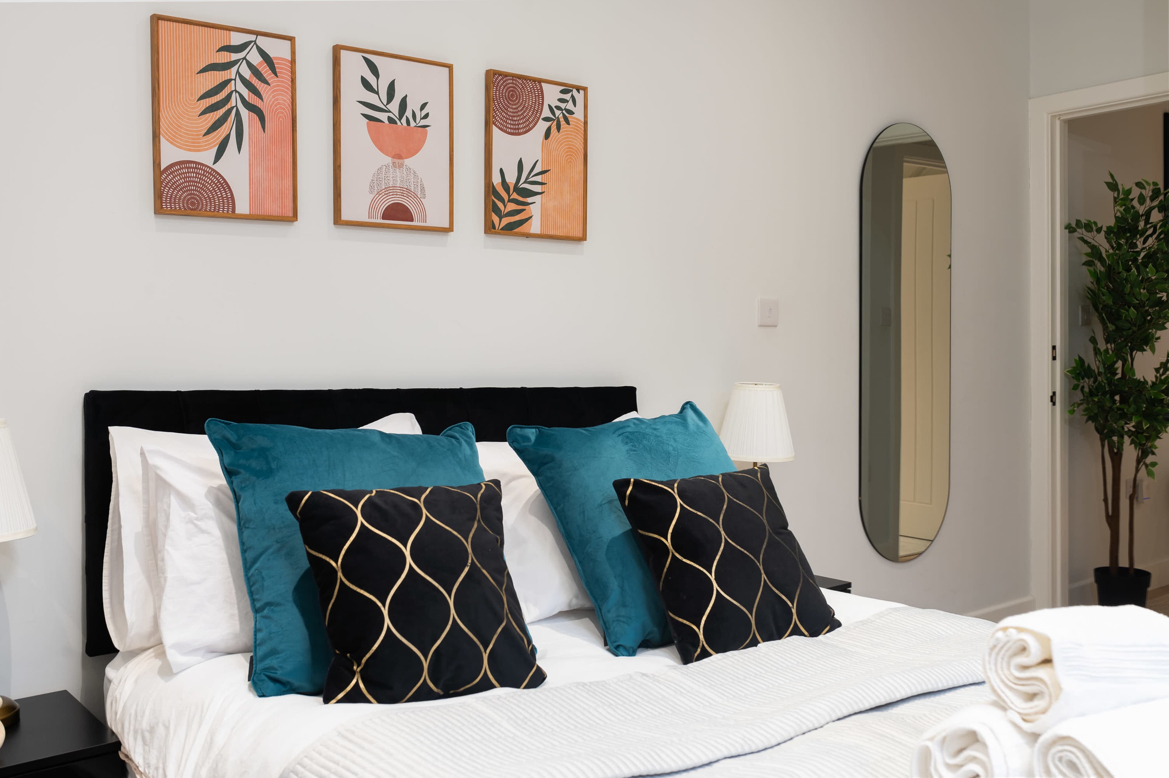 The image shows a neatly made bed with decorative pillows, framed artwork on the wall above, and a mirror beside a plant.