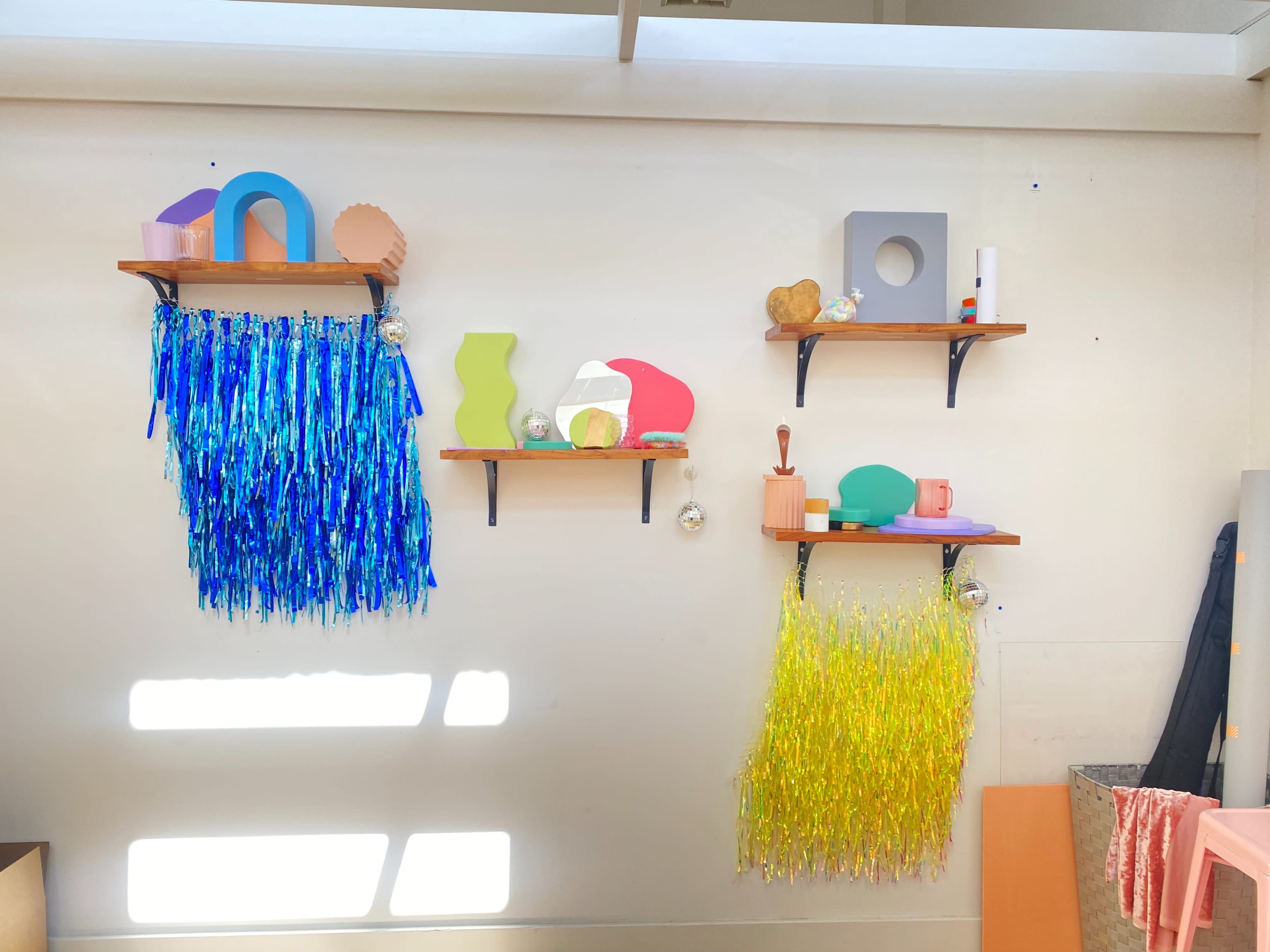 The image displays a white wall with four wooden shelves featuring colorful decorative objects, including shapes and textured materials, with strands of blue and yellow hanging below two of the shelves.