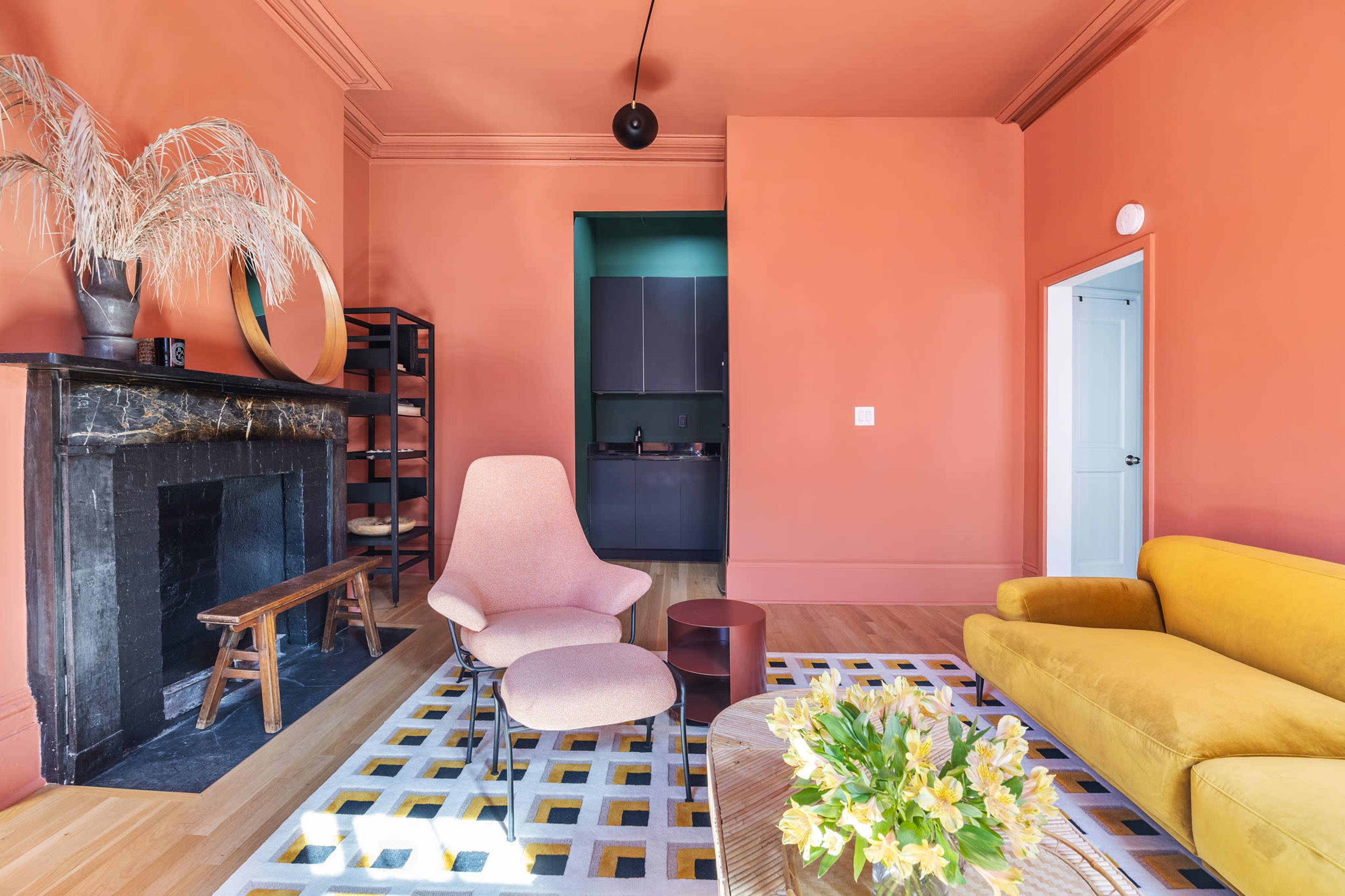 The image shows a stylish interior with an orange-painted room featuring a fireplace, a yellow sofa, a light pink chair, and a patterned rug.