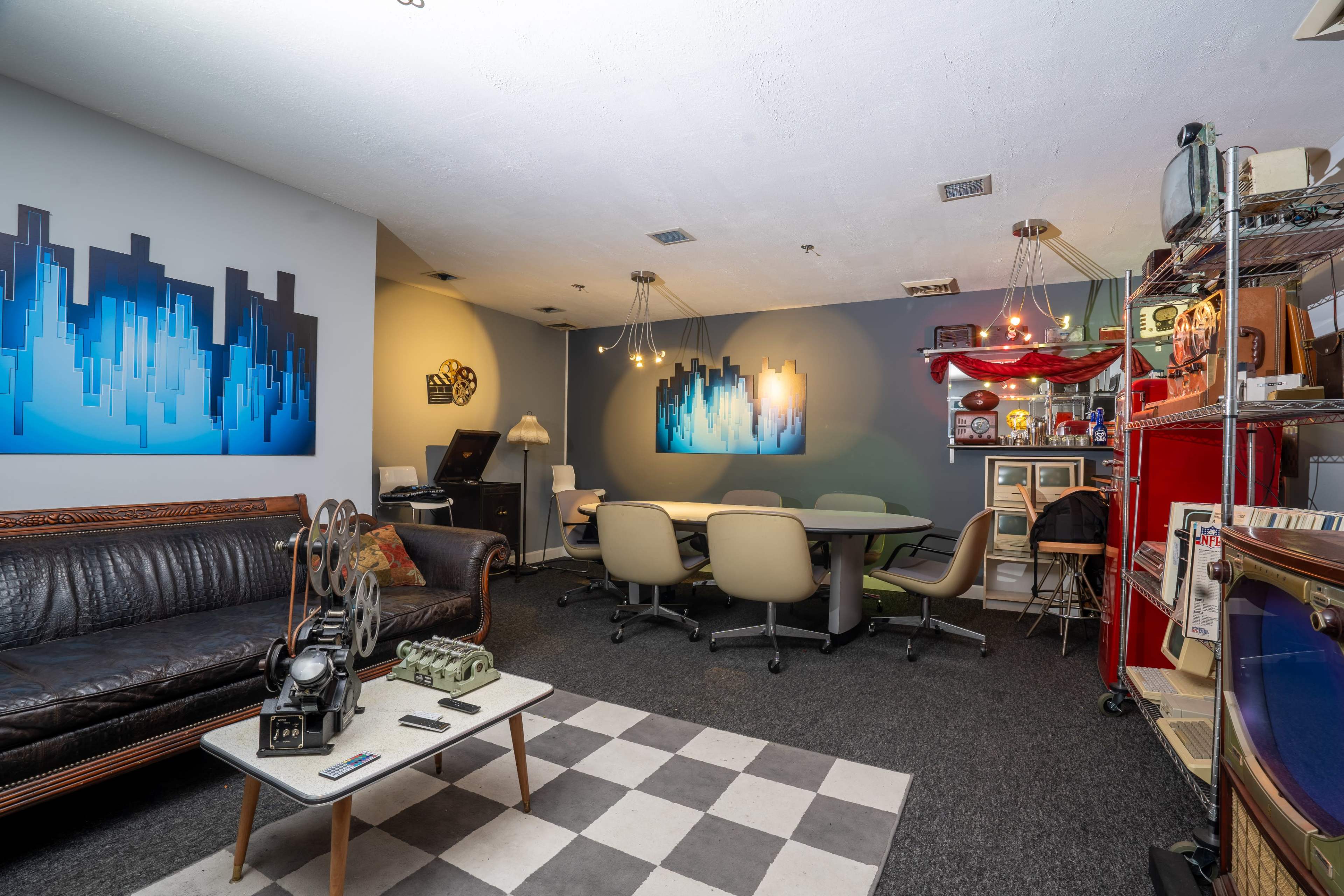 The image shows a creative workspace featuring a checkerboard-patterned floor, a dark leather sofa, a round table surrounded by chairs, and colorful wall art depicting city skylines.