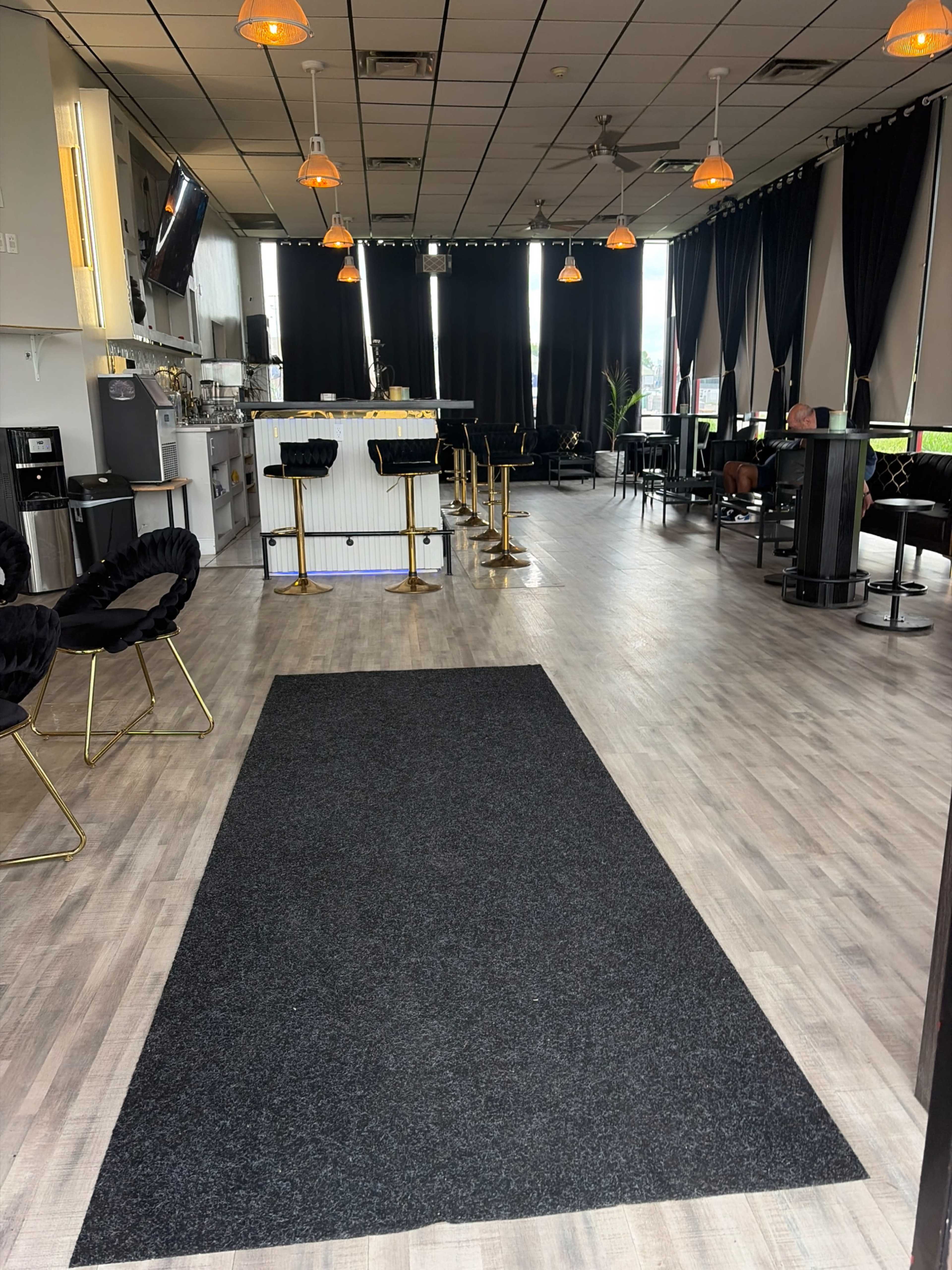 The image shows a modern cafe or lounge with a black carpet runner leading to a bar area, surrounded by seating arrangements and pendant lighting.