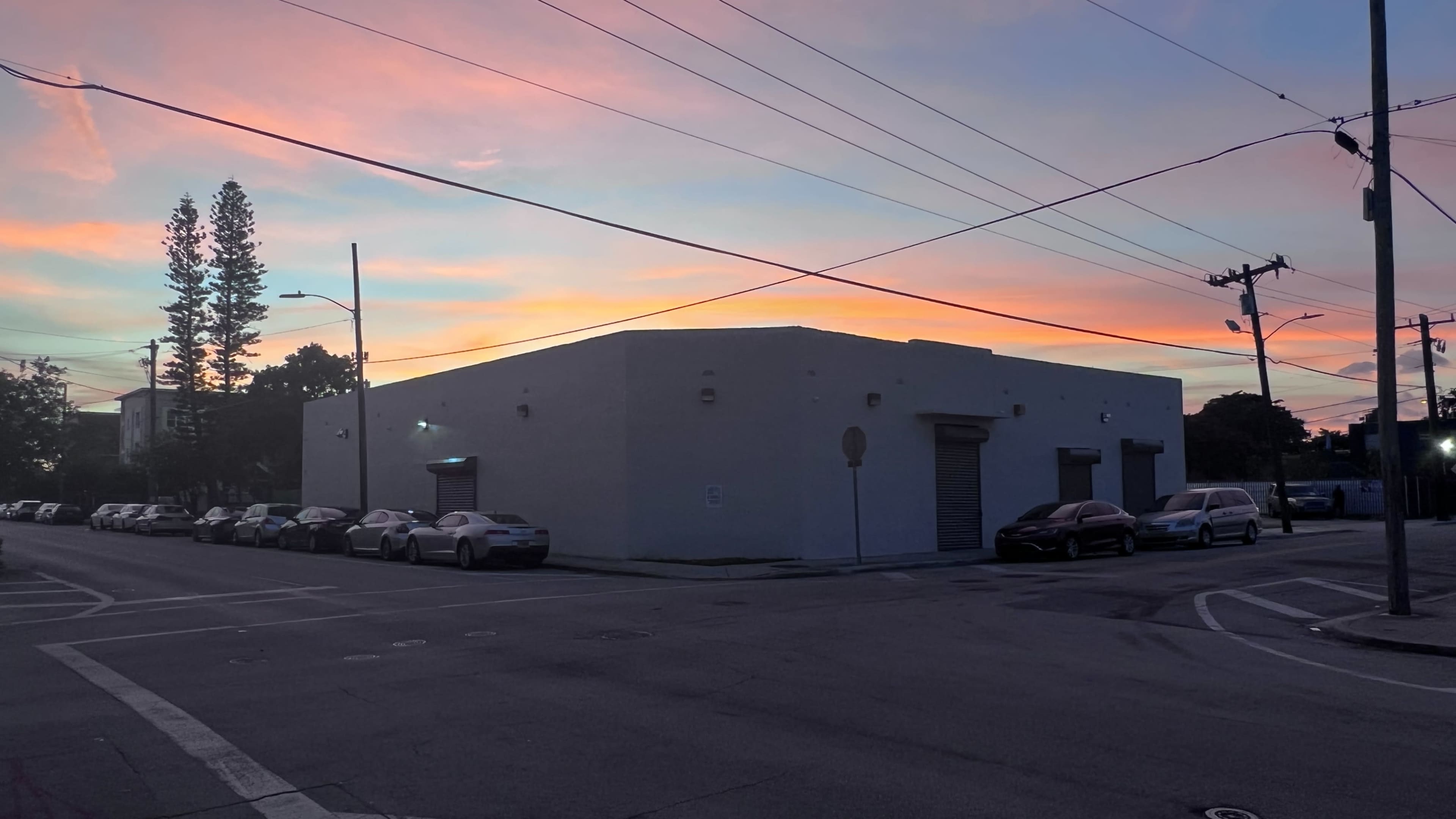 A white building at the corner of a street, with several parked cars and a colorful sky at sunset.