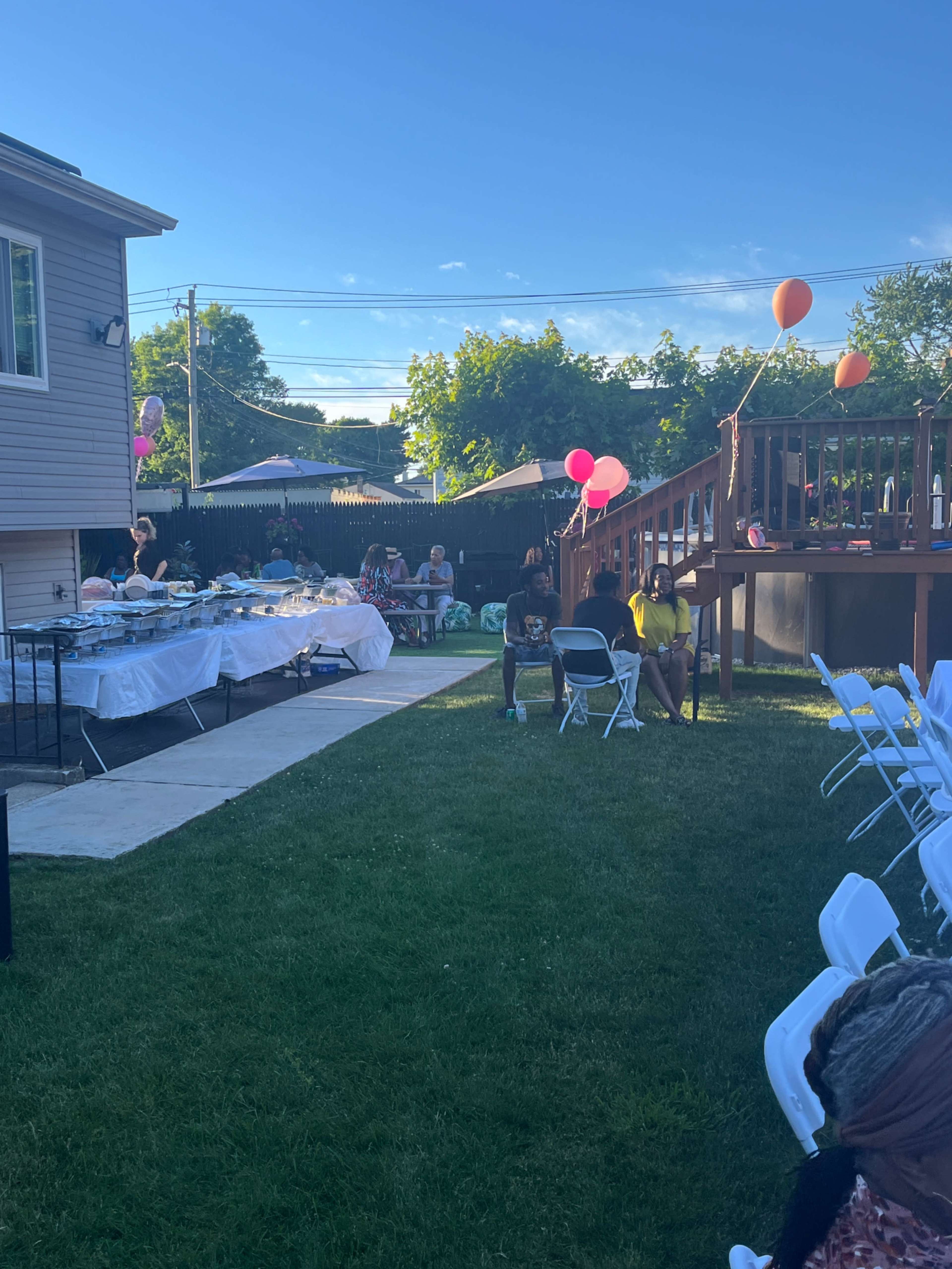 The image shows a backyard party setting with a long table of food, people seated on folding chairs, and balloons in the background.
