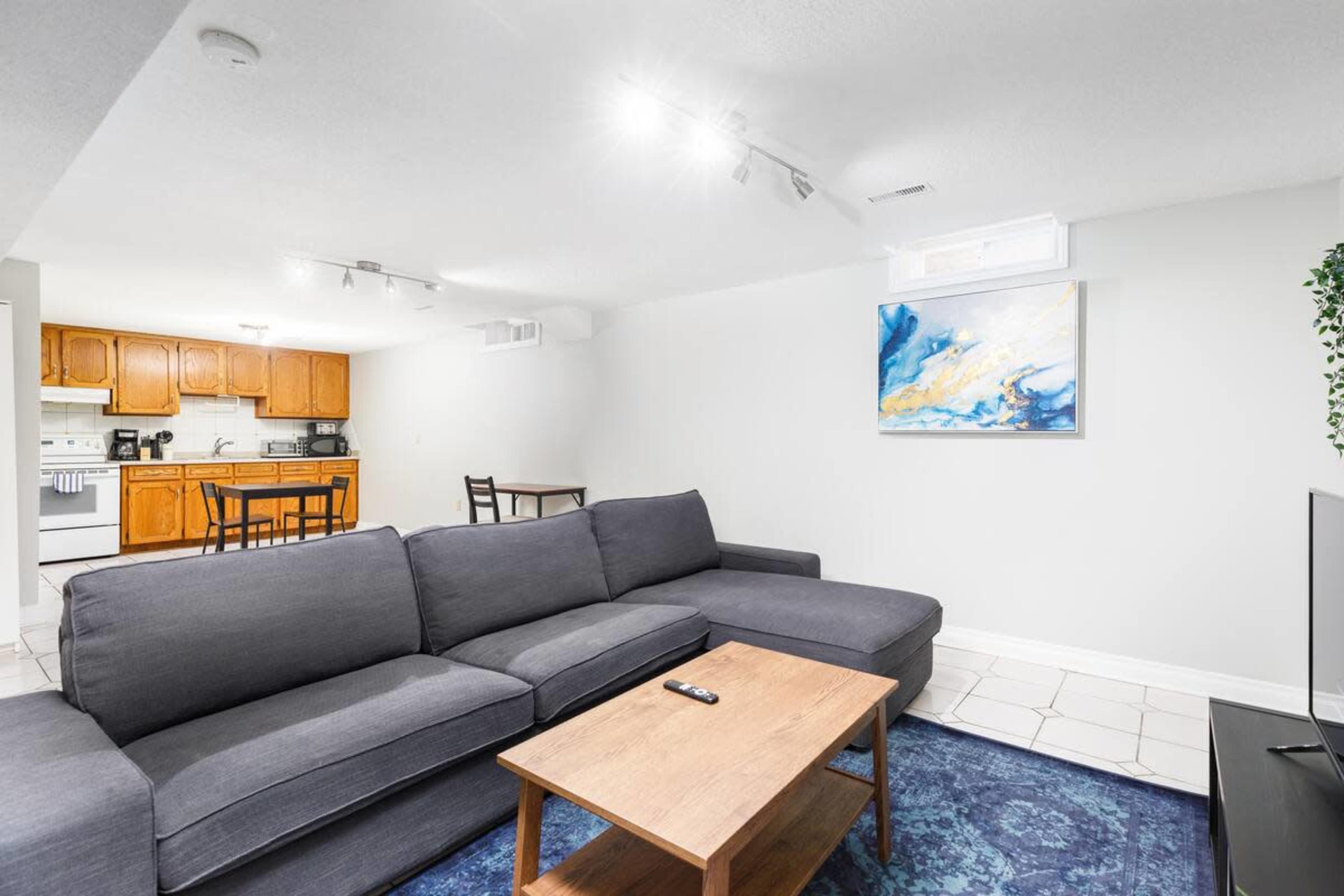 A modern living area with a grey sectional sofa, a wooden coffee table, and a kitchen in the background featuring wooden cabinets and a dining table.