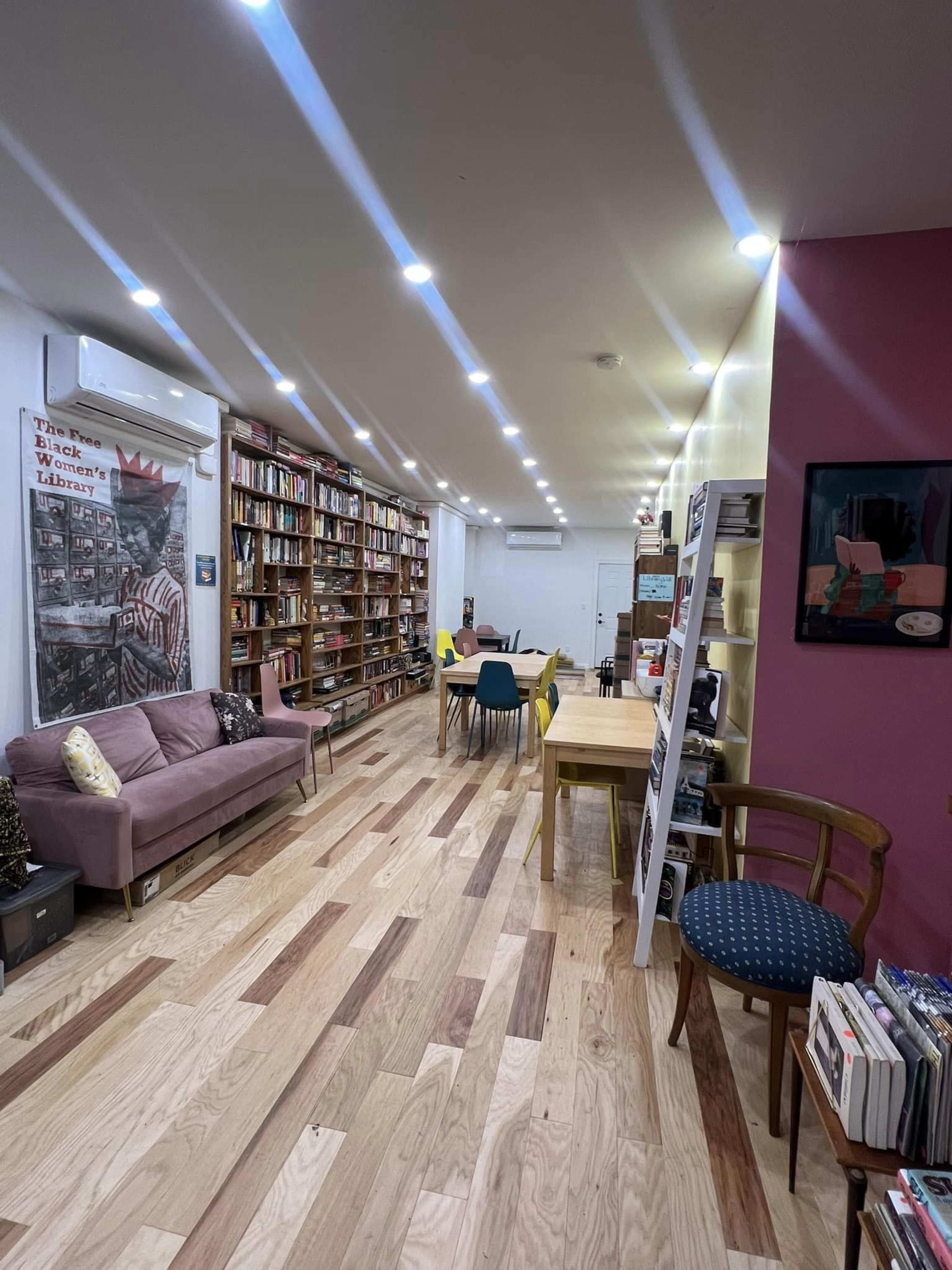 A library or reading space with wooden floors, shelves filled with books along the walls, and tables and chairs arranged for seating.