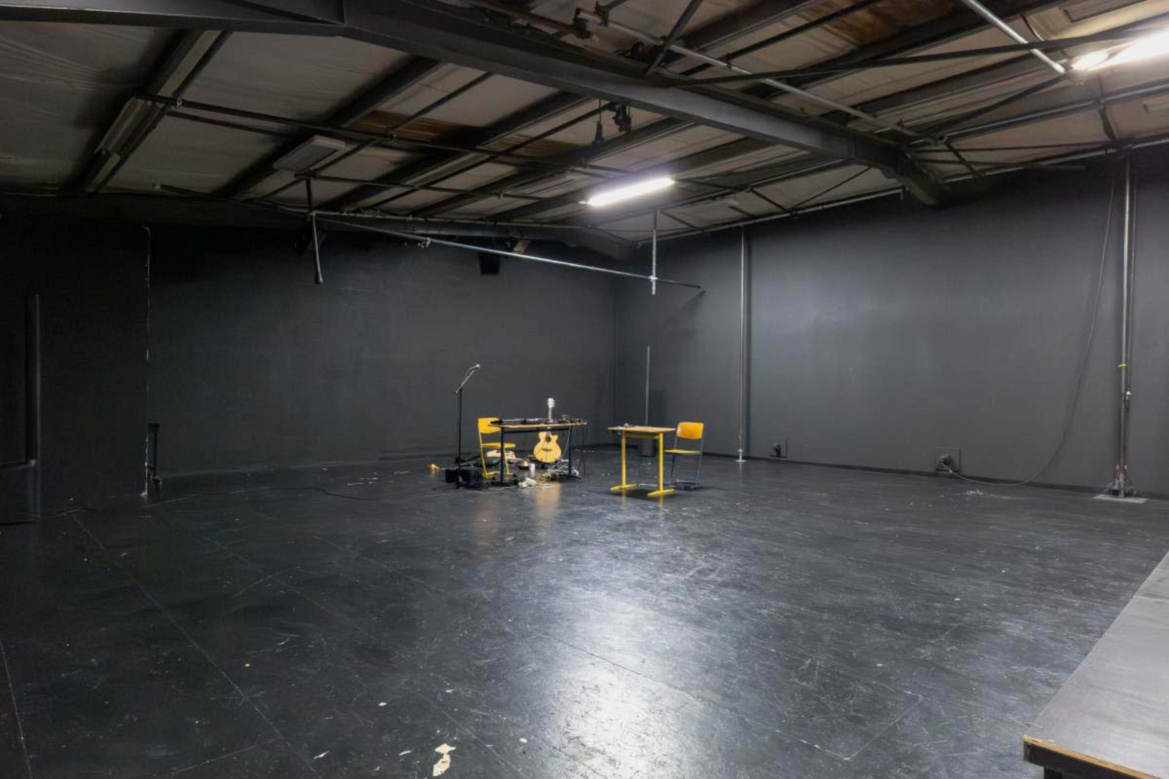The image shows a spacious, dimly lit room with black walls and a bare floor, featuring a table, two chairs, a microphone, and a guitar on a stand.