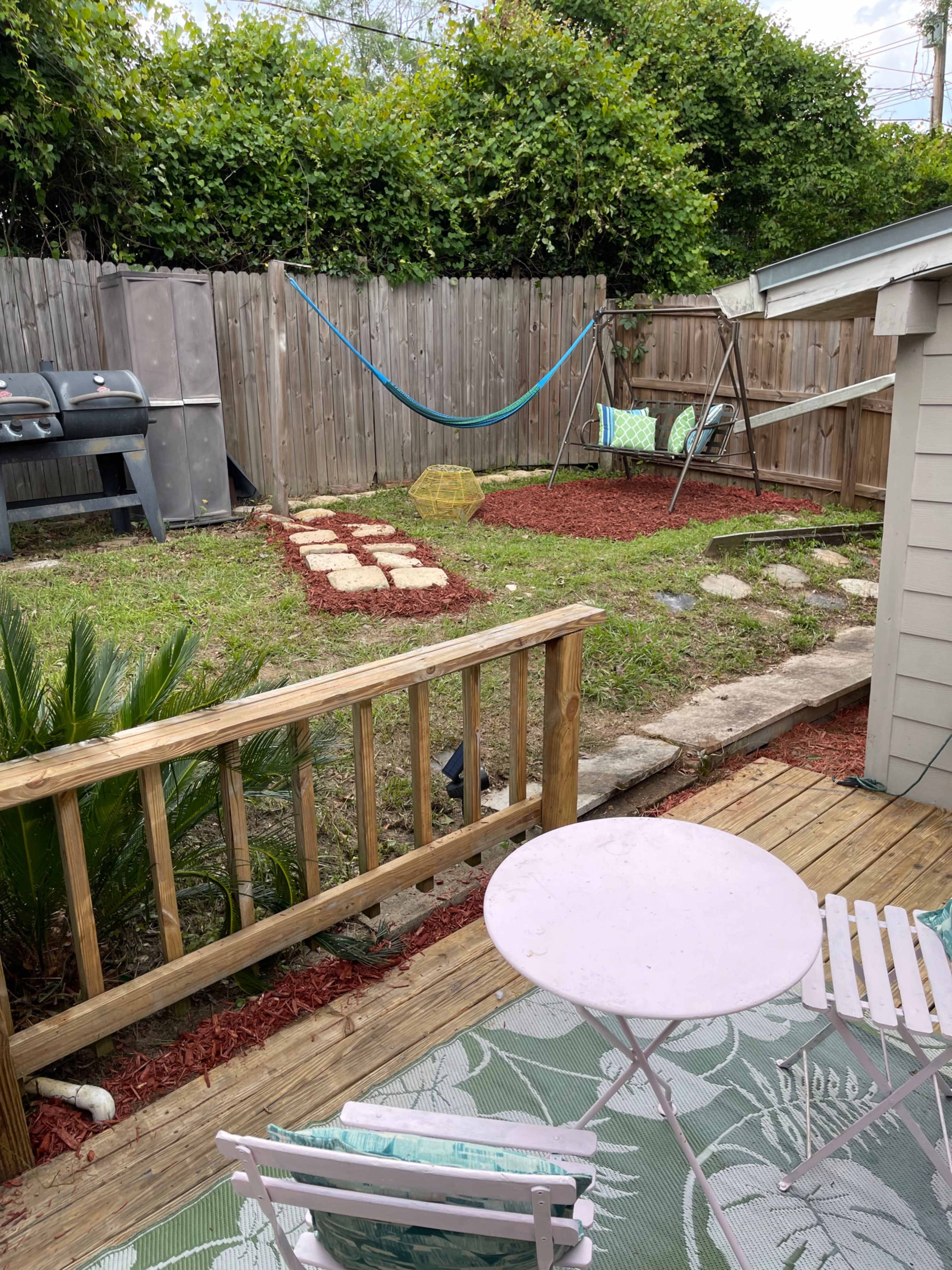 A backyard with a wooden deck, a small table and chairs, a grass area with stepping stones, a hammock, a swing set, and a grill against a wooden fence.