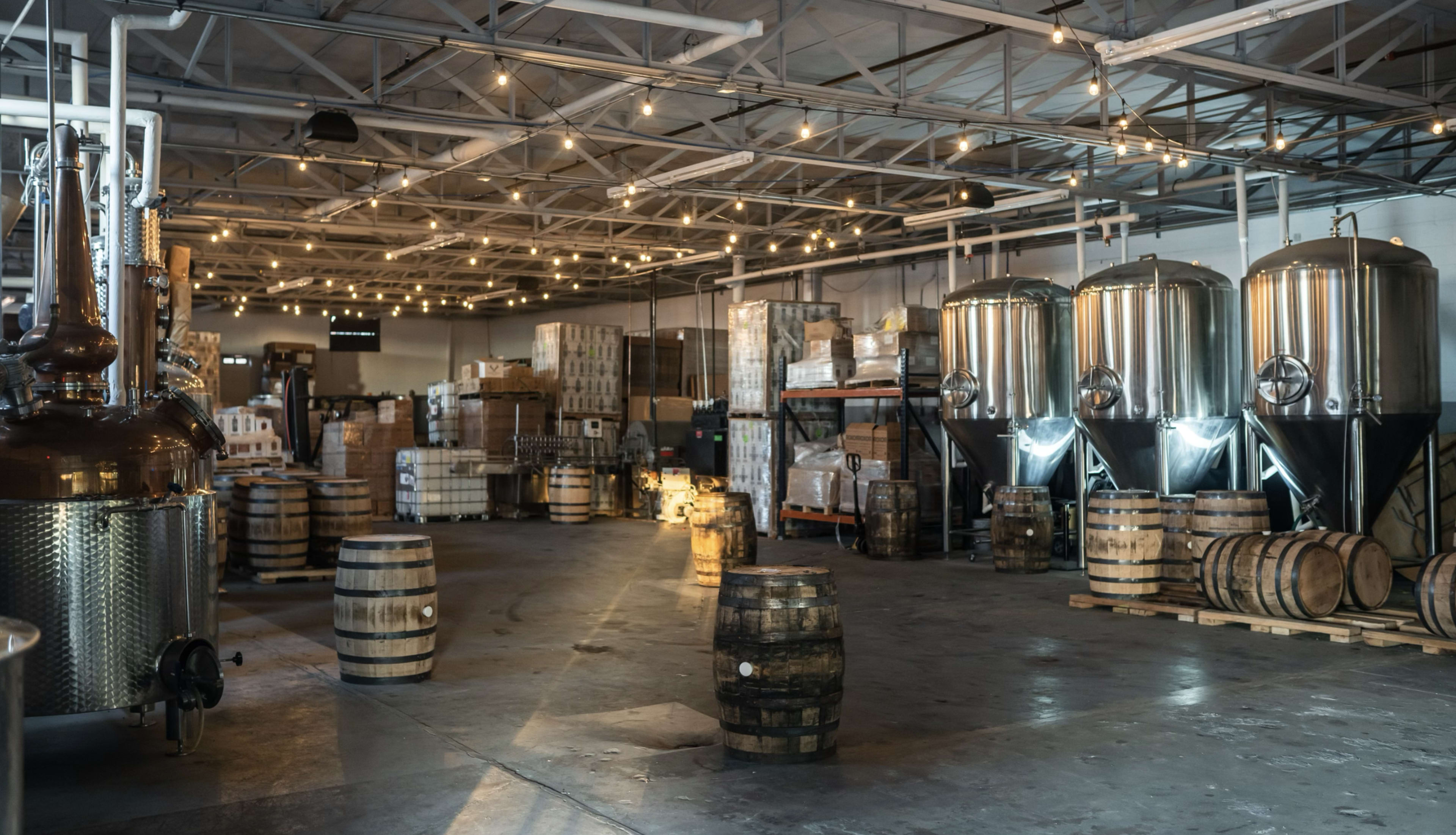 The image shows a spacious warehouse with large metal fermentation tanks, wooden barrels, and stacks of boxes, all under warm, overhead string lights.