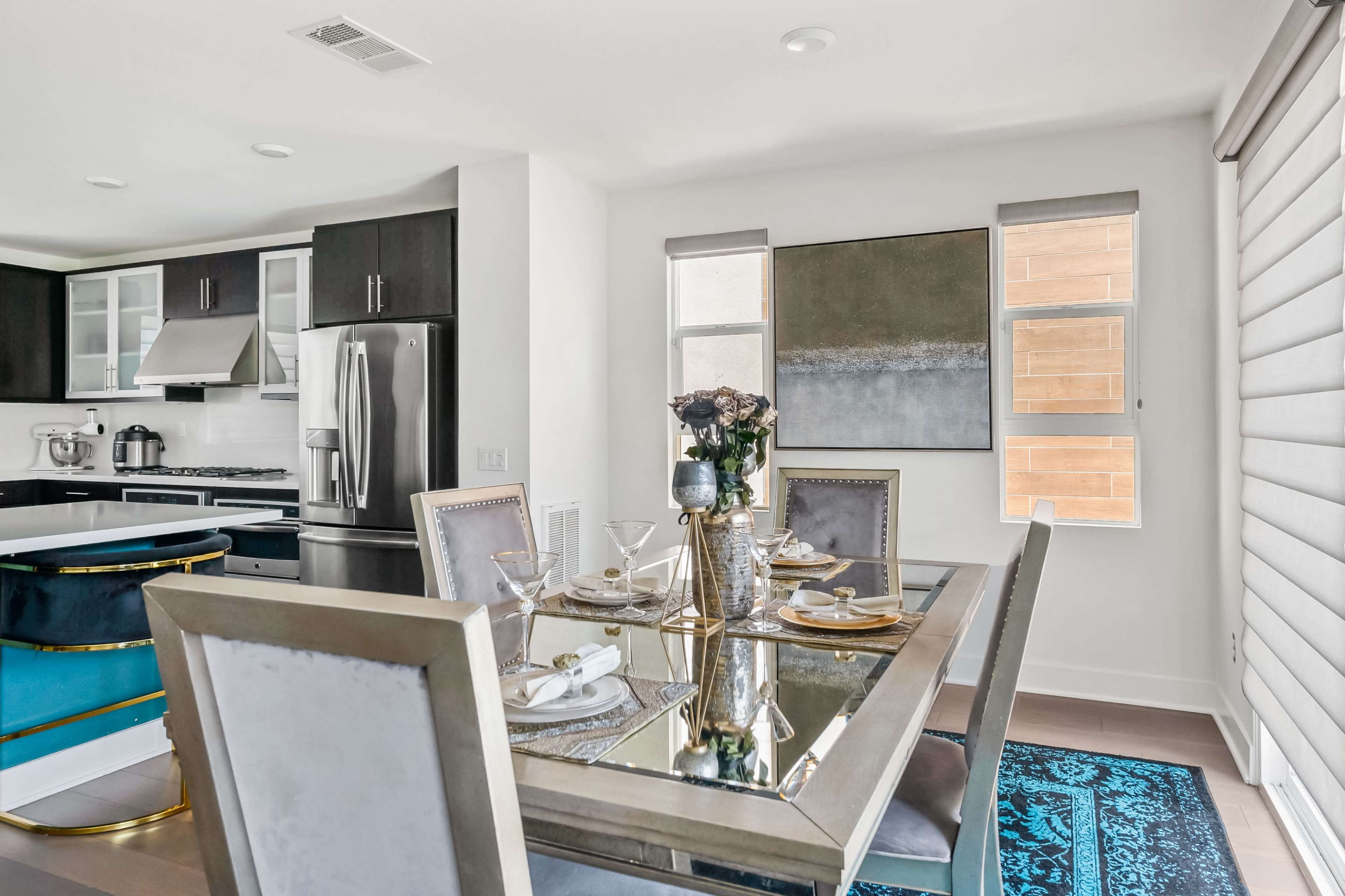 A modern dining area features a glass-topped table set for four, surrounded by upholstered chairs, with a kitchen visible in the background.