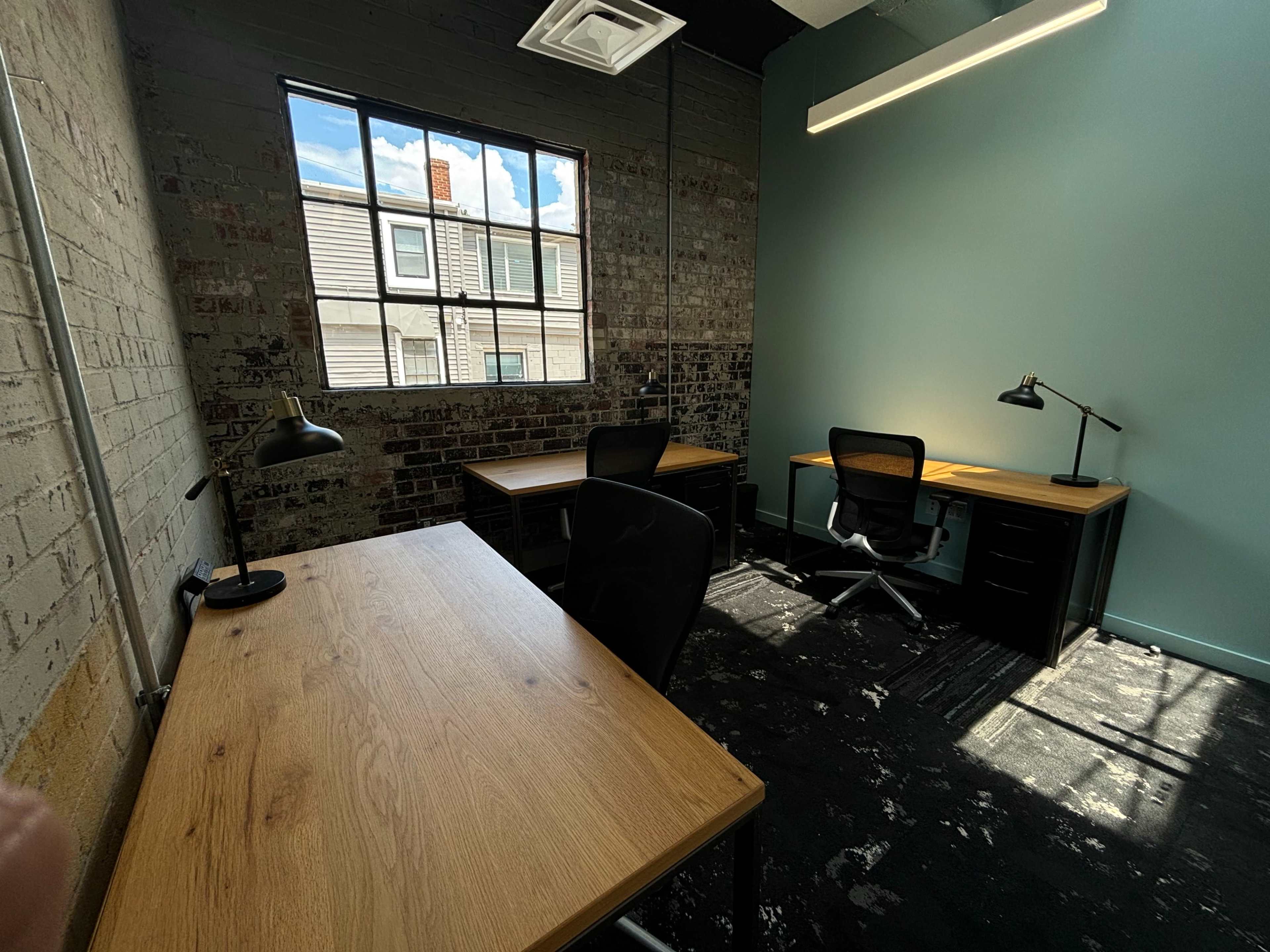 A well-lit office space with two wooden desks and a chair, featuring exposed brick walls and a large window that opens to an exterior view.