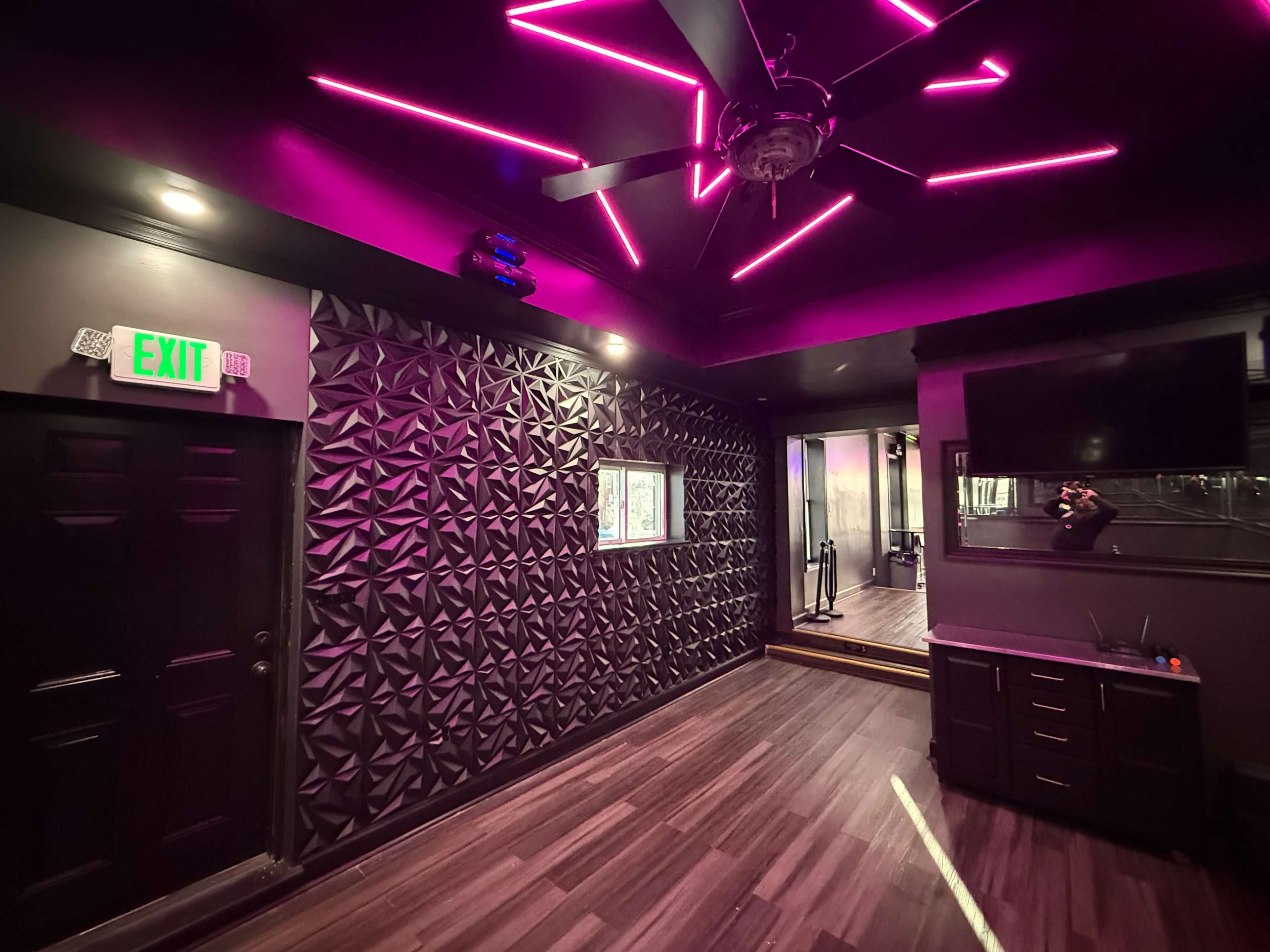 The image shows a modern interior room with a textured black wall, pink neon lighting on the ceiling, and wooden flooring.
