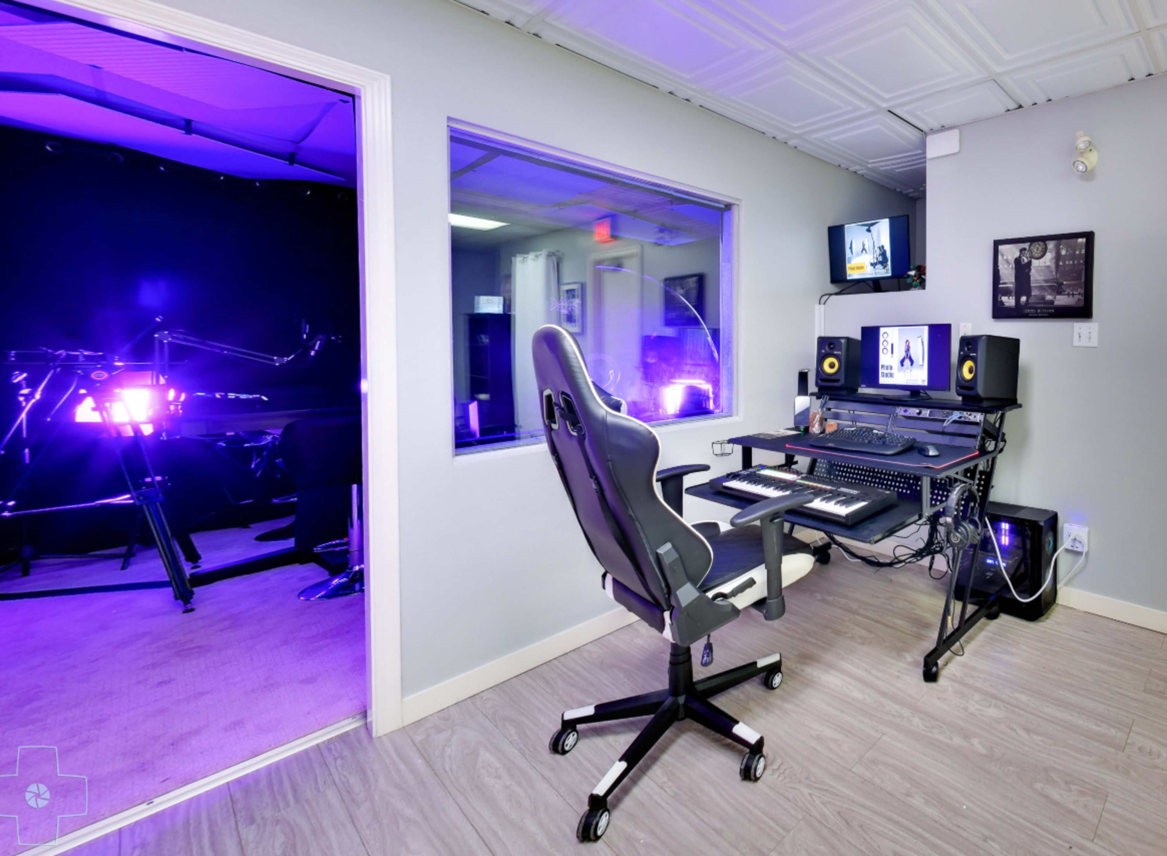 A music production studio with a desk featuring monitors, a keyboard, and audio equipment, adjacent to a room illuminated with purple lighting.